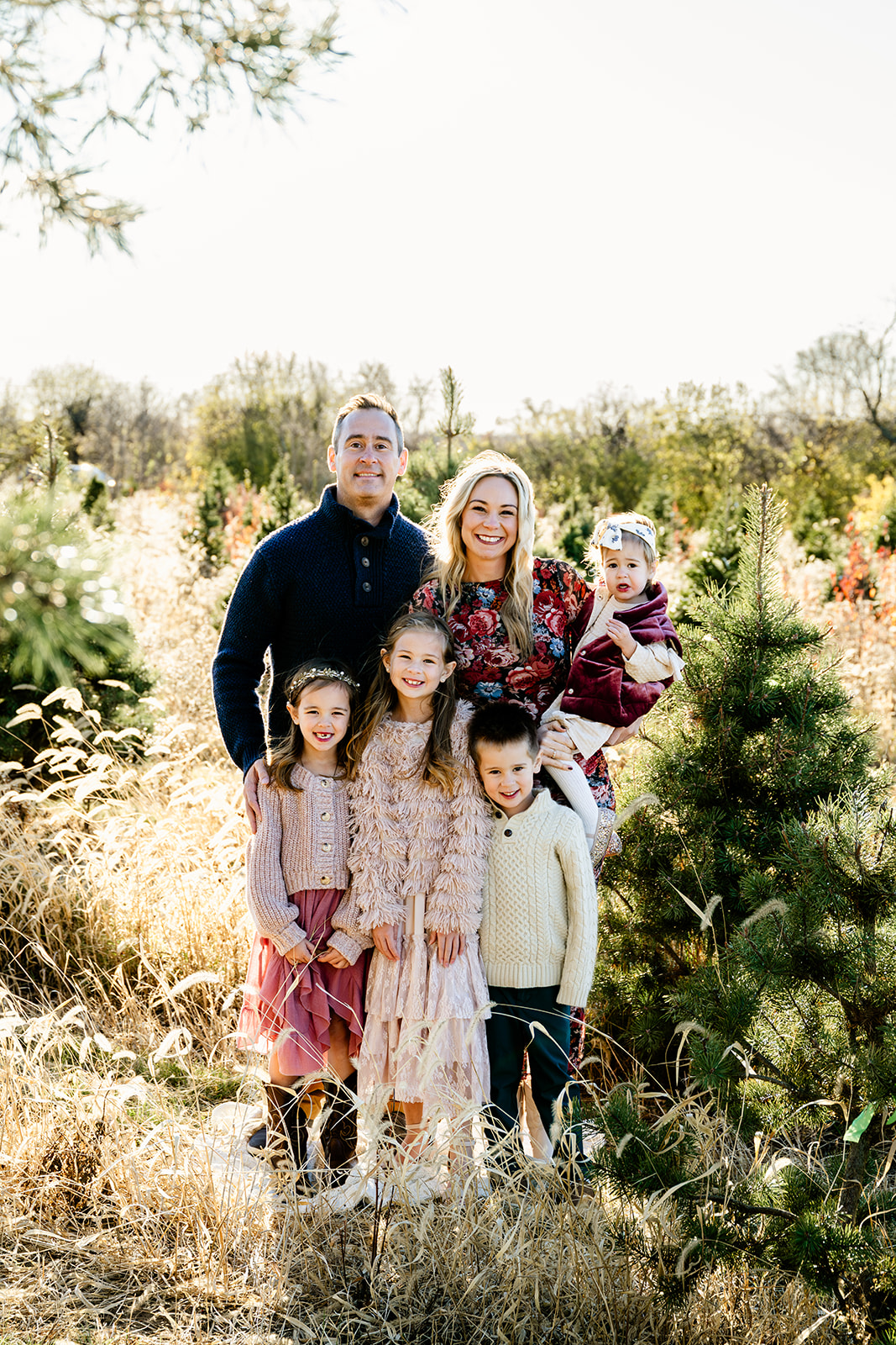 A happy family of 6 stands in a christmas tree farm at sunset smiling