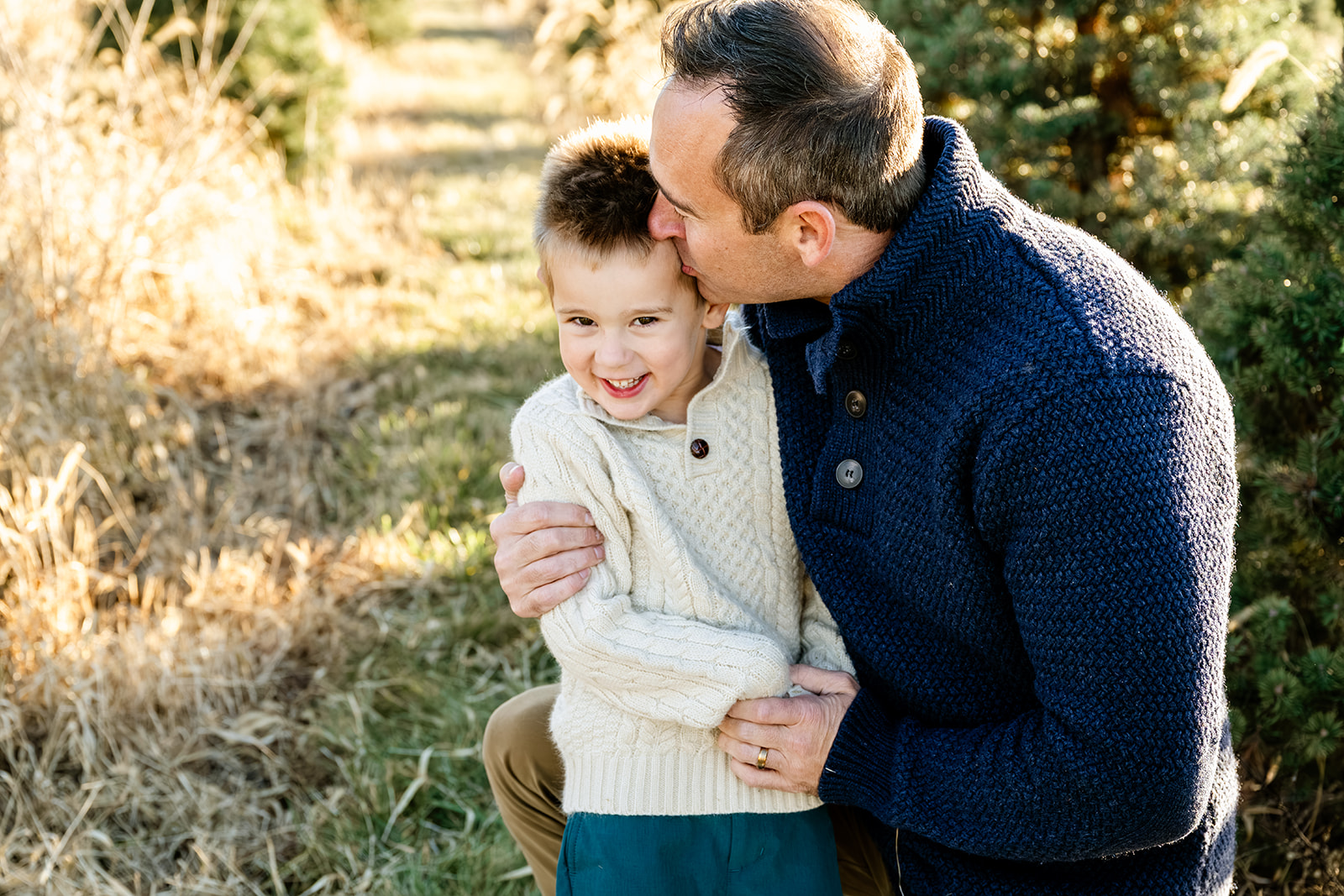 A happy toddler in a white sweater is kissed by dad in a blue sweater in a park trail while staying at kid-friendly hotels in Chicago