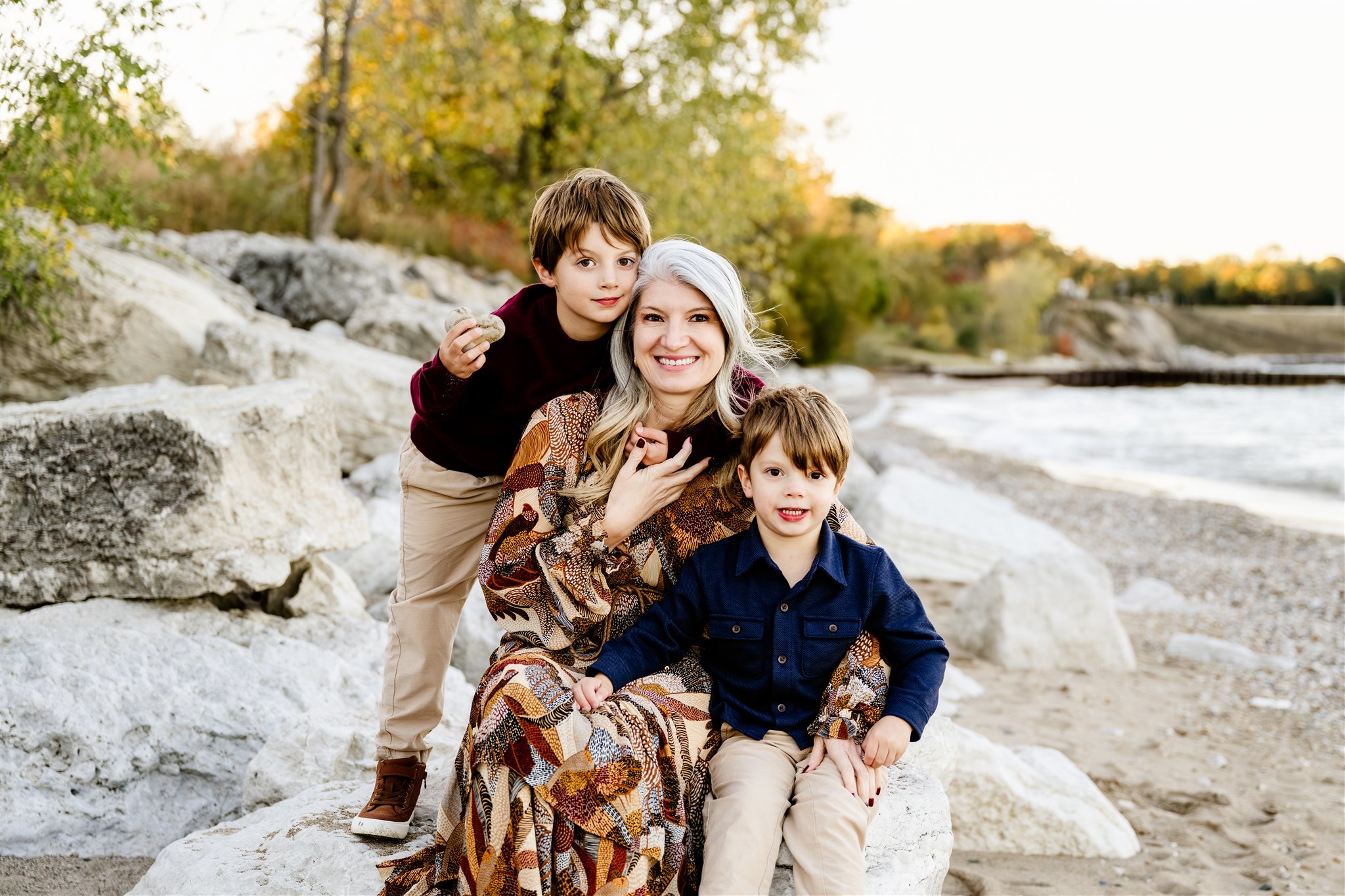 Two toddler boys hug and smile with their mom on a boulder on a beach at sunset