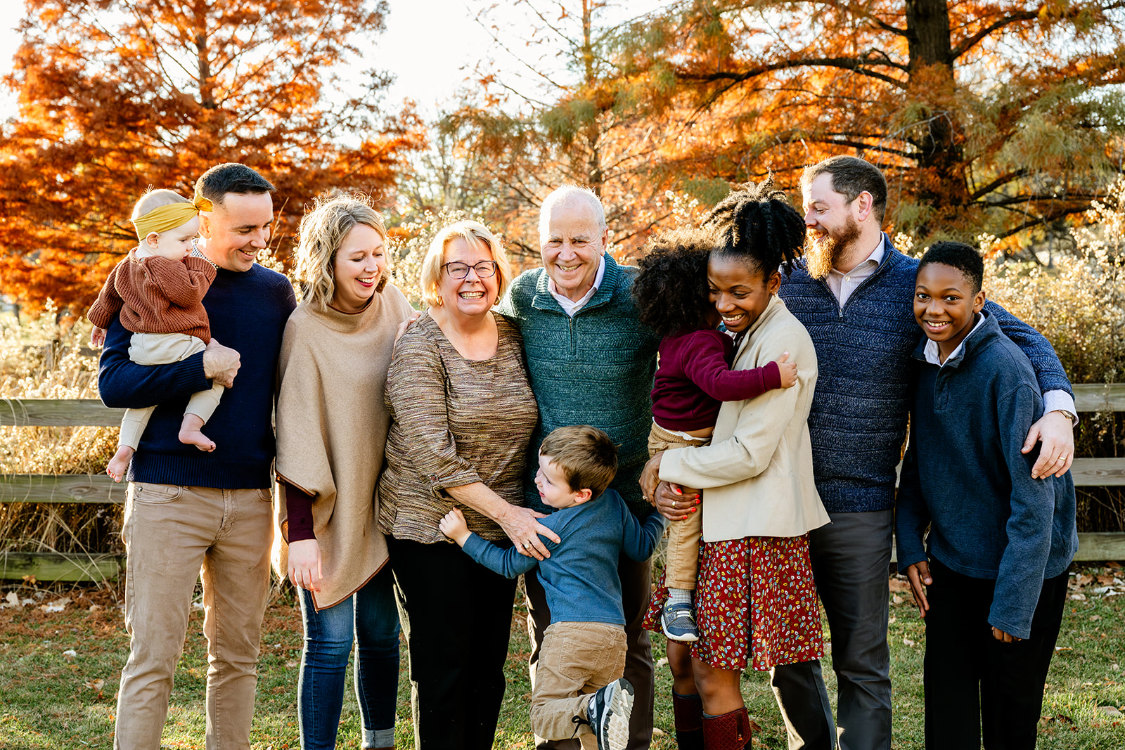 A large extended family laughs and hugs while standing in a park in fall wearing sweaters