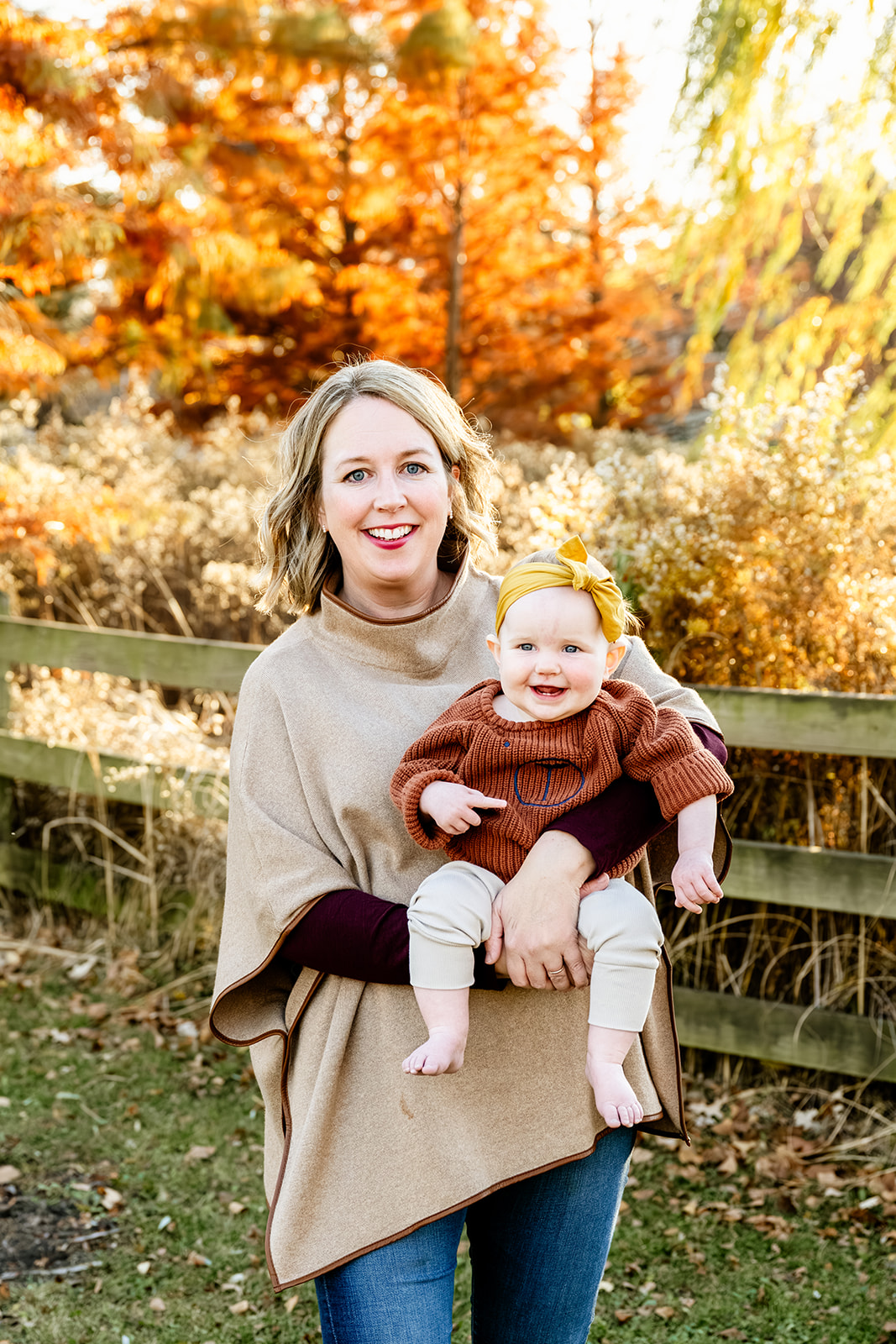 A smiling mother stands in a park in fall in a tan sweater holding her baby daughter in a brown sweater after loving North Shore Nannies