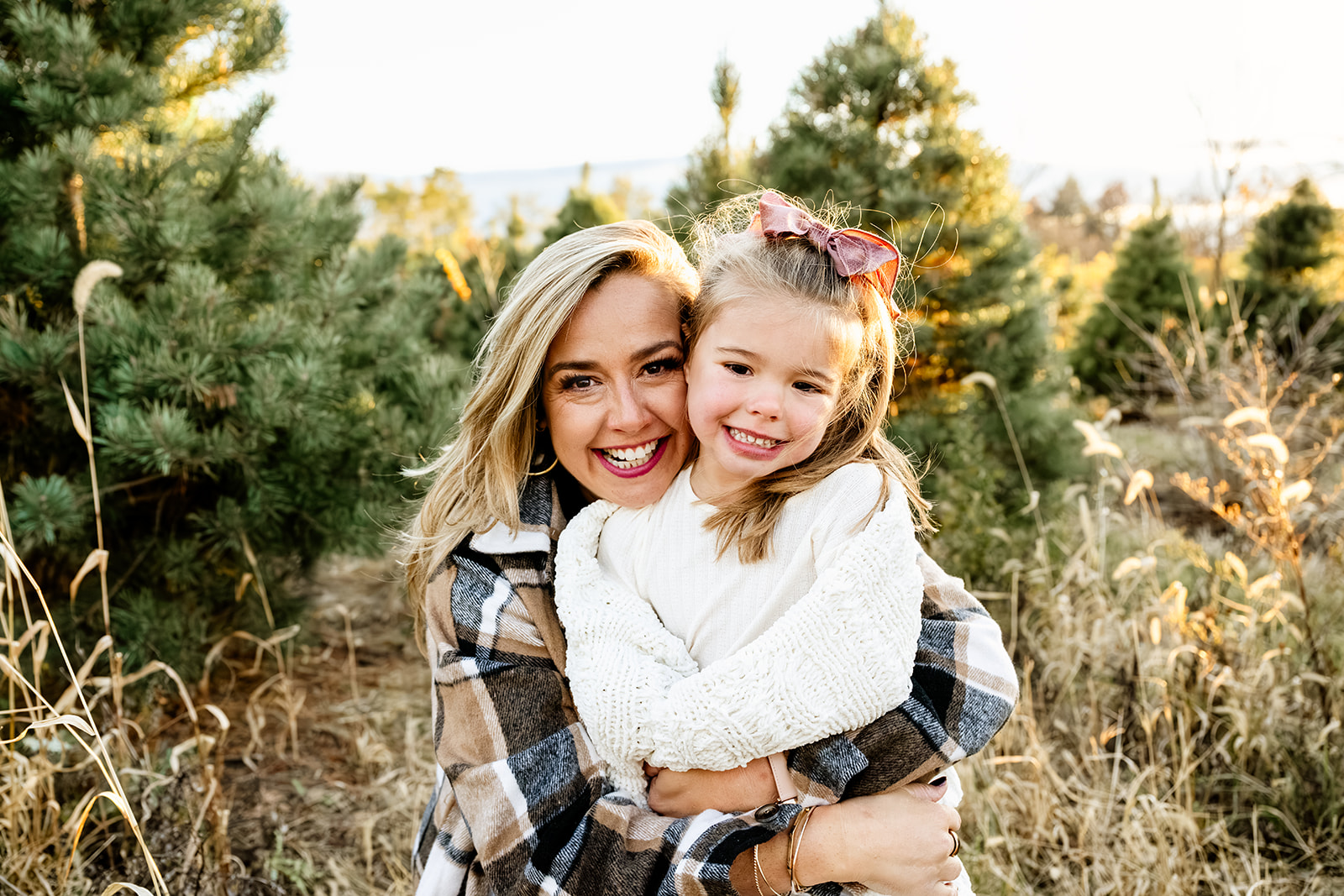 A happy mom in plaid hugs her toddler daughter in a christmas tree farm at sunset