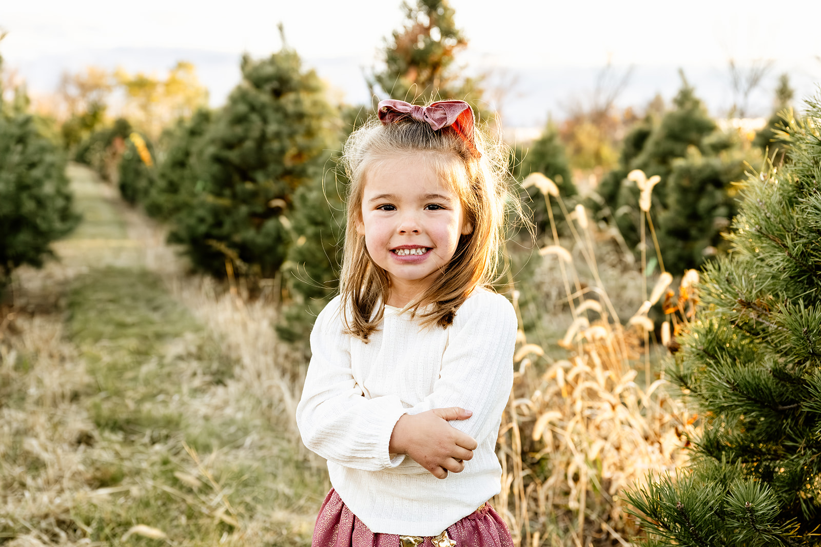 A toddler girl in a white shirt crosses her arms while smiling in a christmas tree farm at sunset after some speech therapy in Naperville