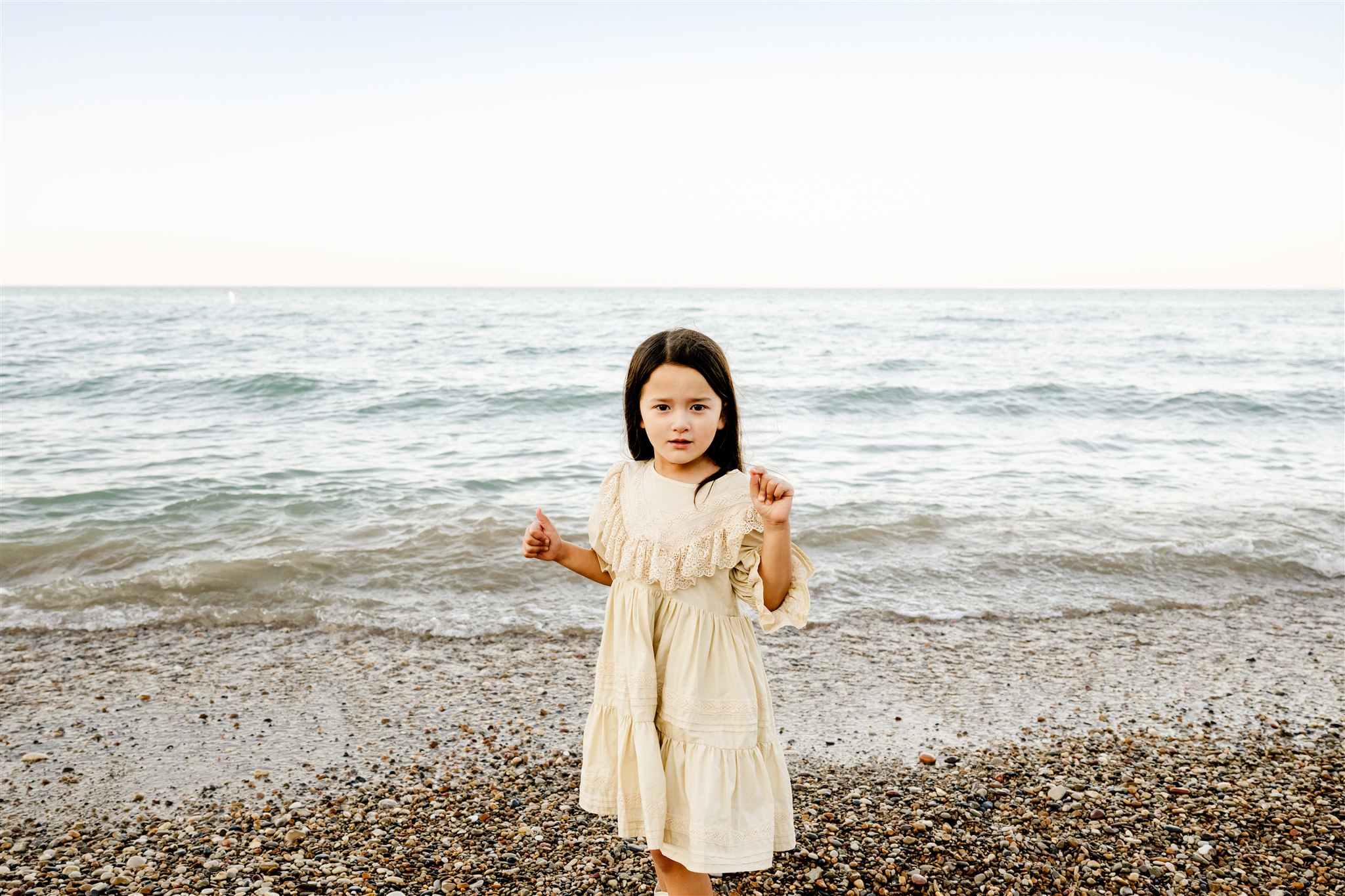 A toddler girl in a cream dress dances on a beach at sunset