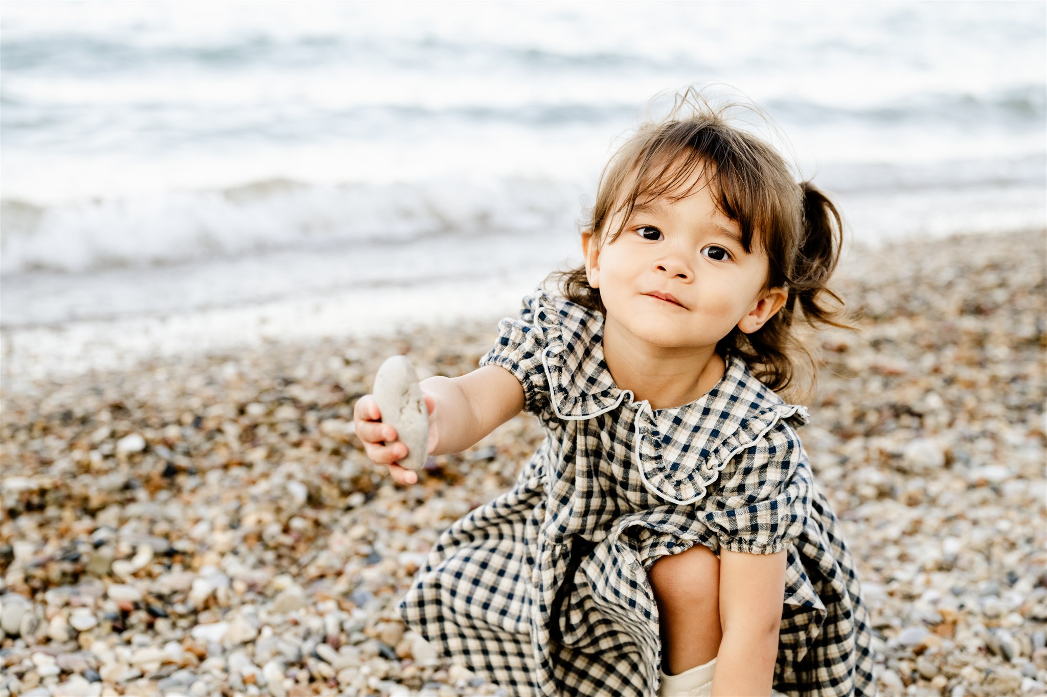 A toddler girl in a plaid dress shoes off a rock on the beach before visiting splash pads in Naperville