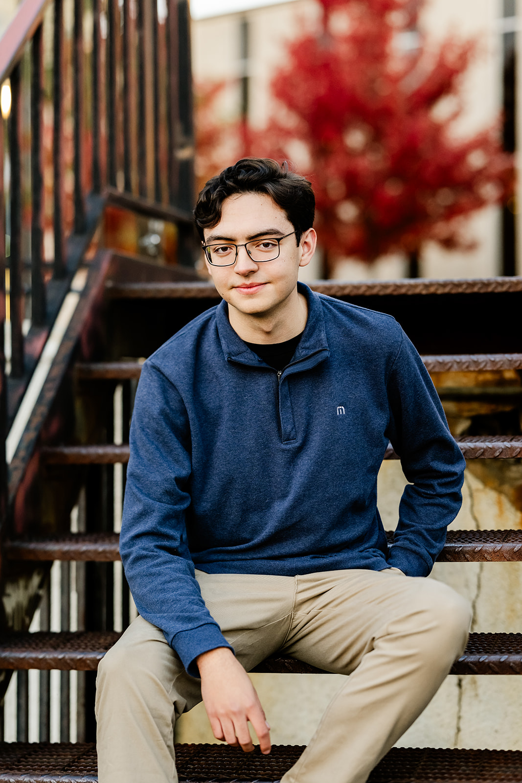 A graduate in a blue sweater and khakis sits on a metal staircase in an alley smiling