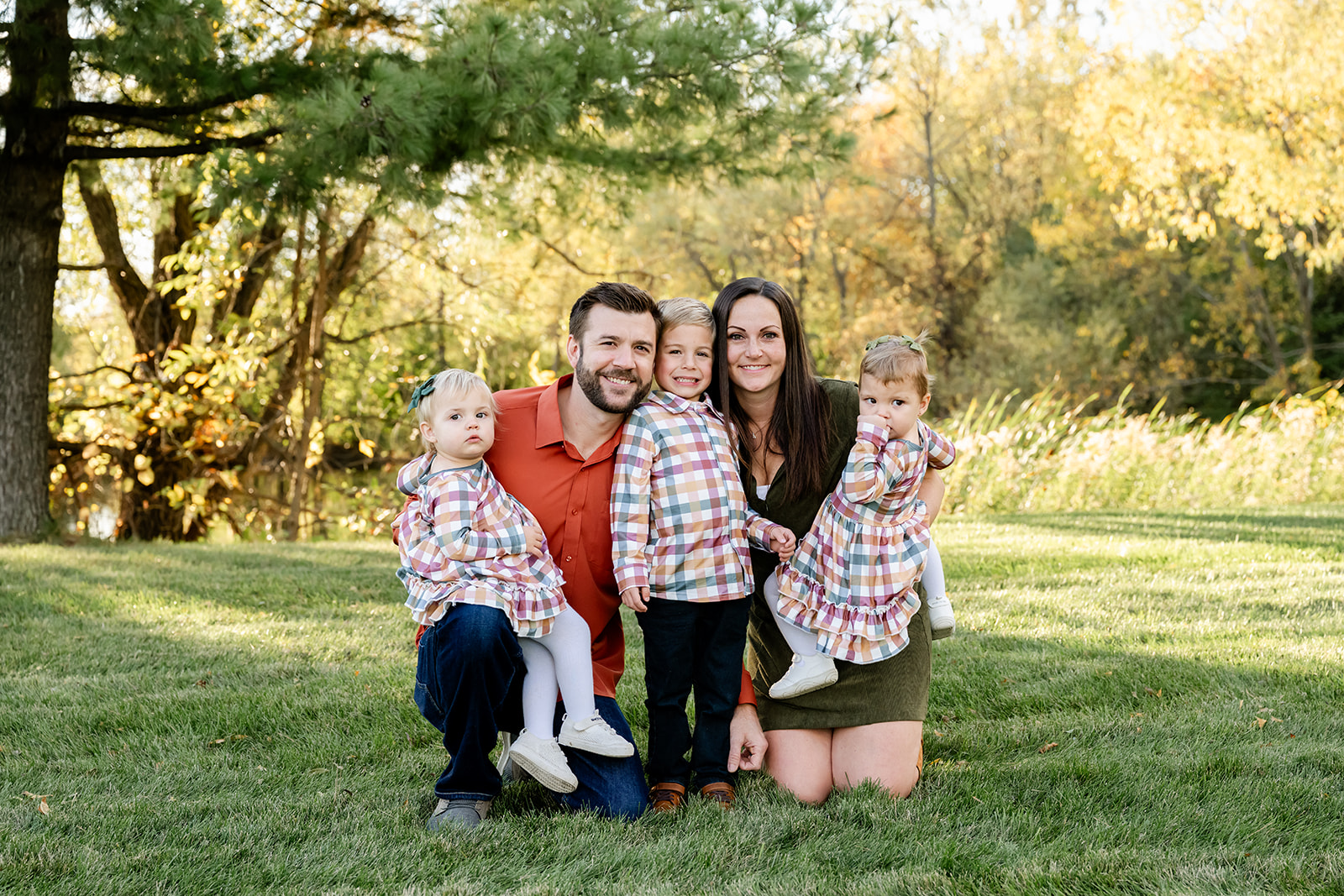 Mom and dad kneel in a park lawn with their three toddlers in matching plaid