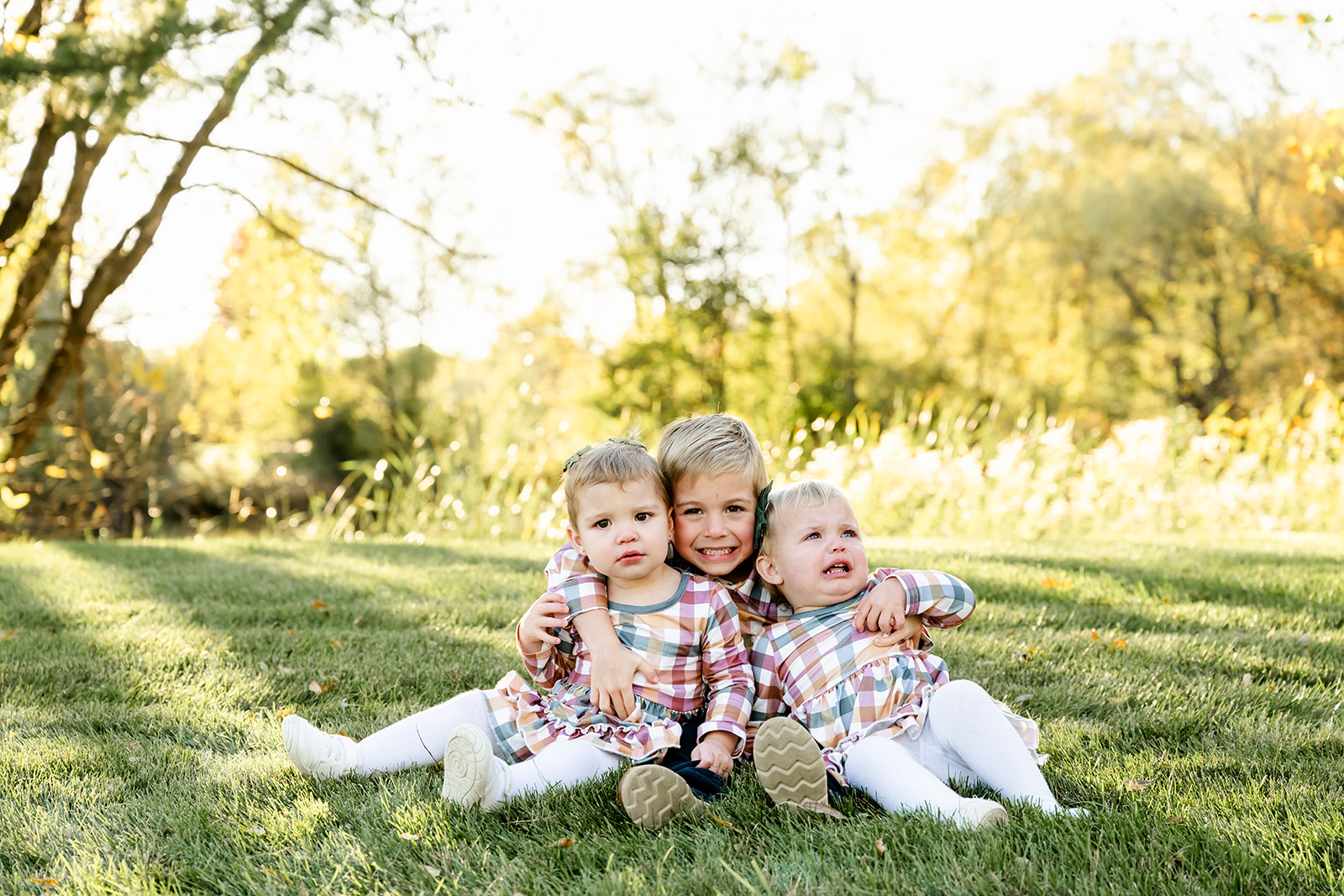 A toddler boy reaches over hugging his baby sisters in matching plaid dresses in a park lawn after visiting daycares in North Shore