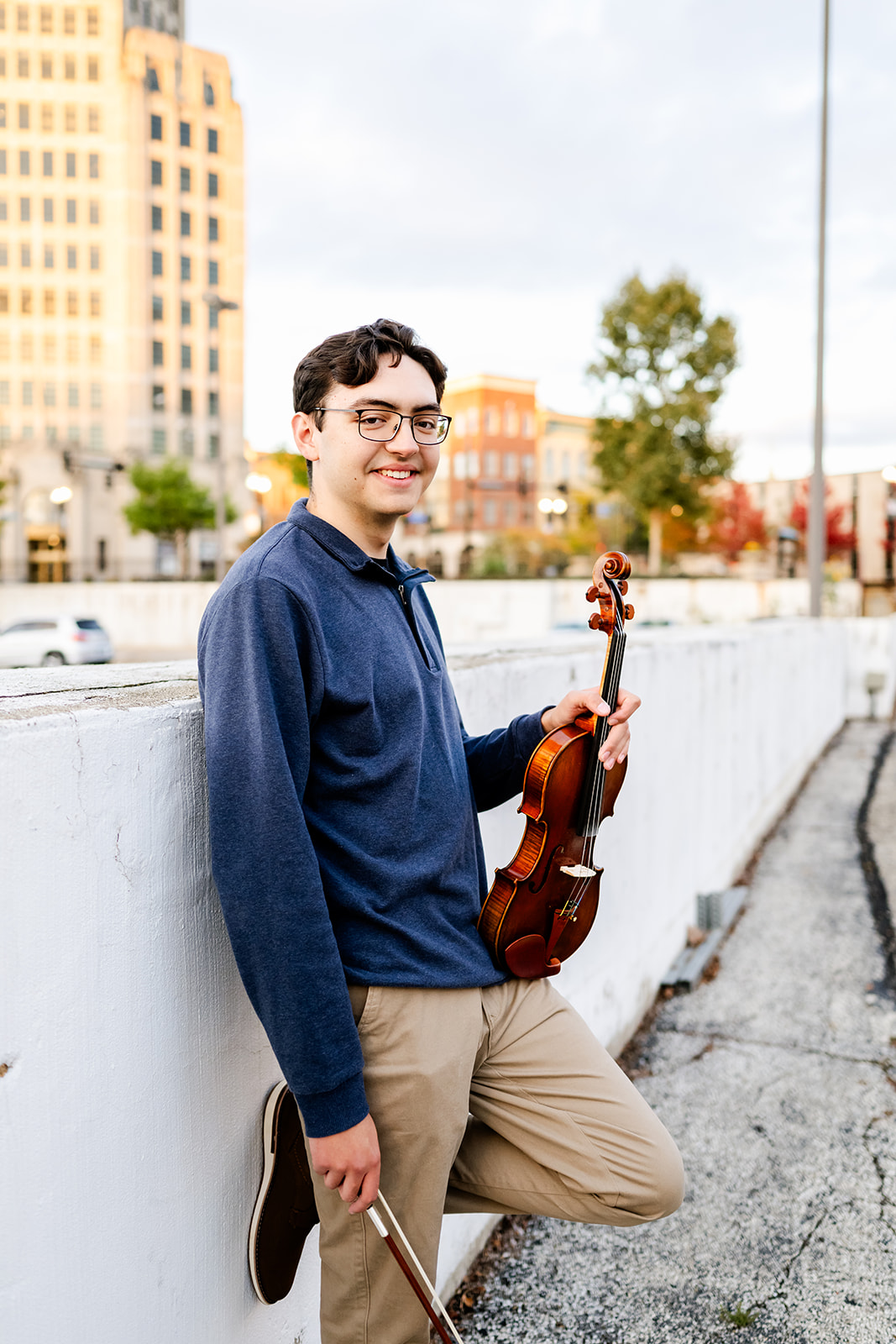 A teenager leans against a concrete wall with his violin at sunset