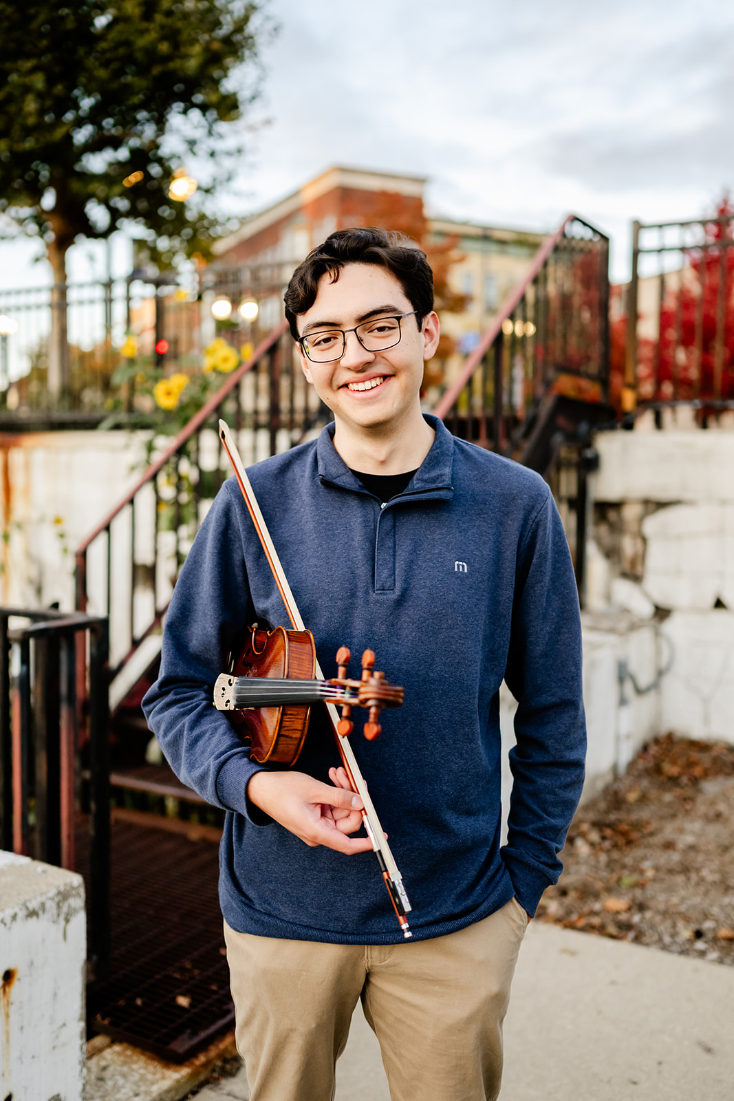 A happy high school senior in a blue sweater stands in a garden holding his violin under his arm after exploring graduation party venues in Chicago