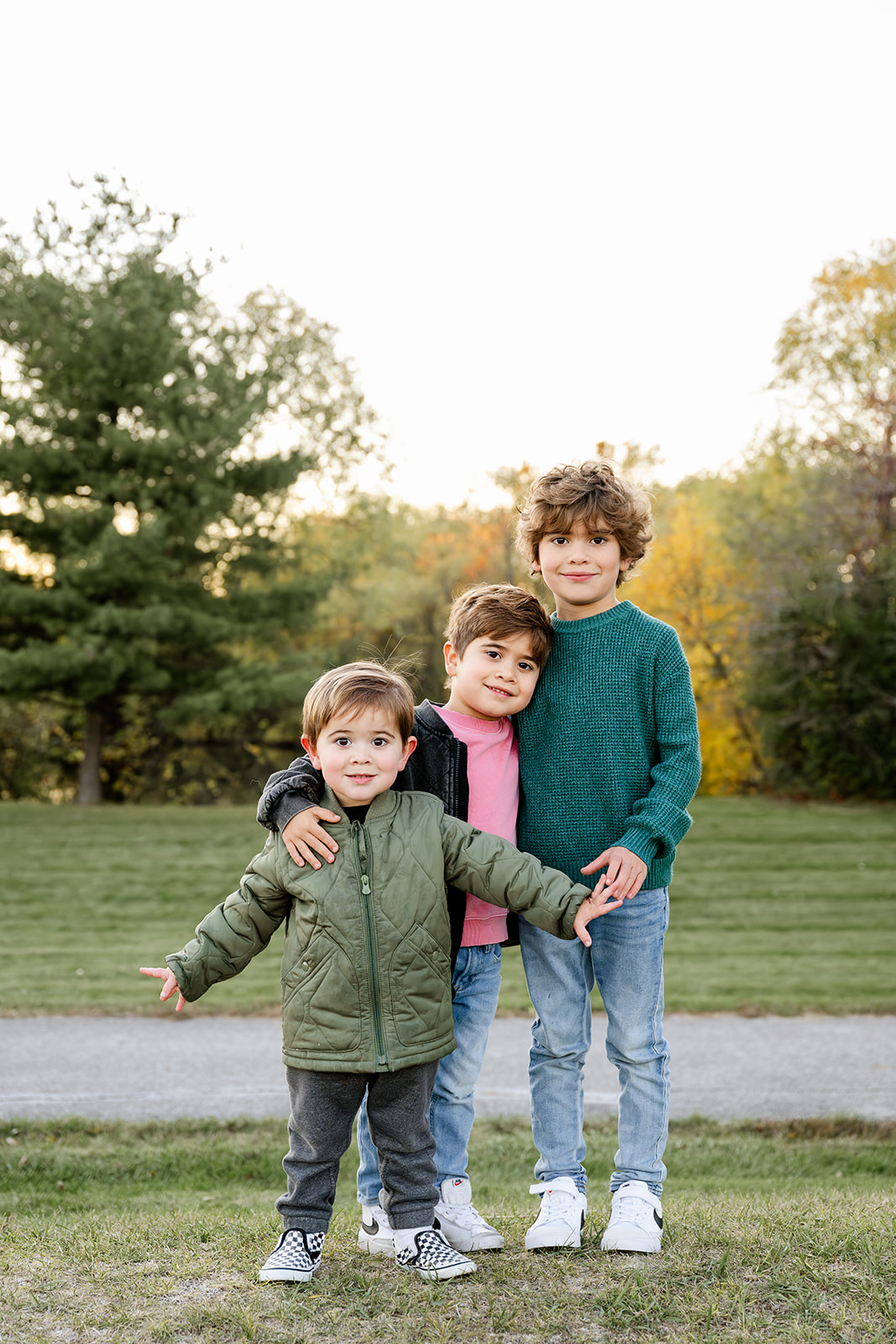 Three toddler brothers stand in a park lawn at sunset with smiles and arms around each other
