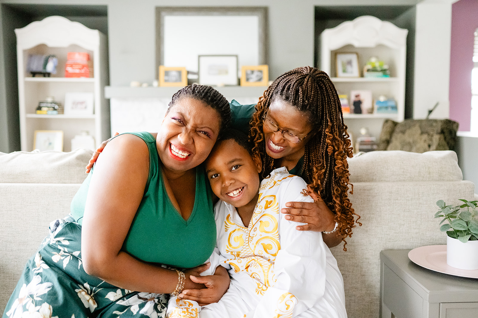 Smiling moms in green hug and smother their young son in white on a couch in a living room