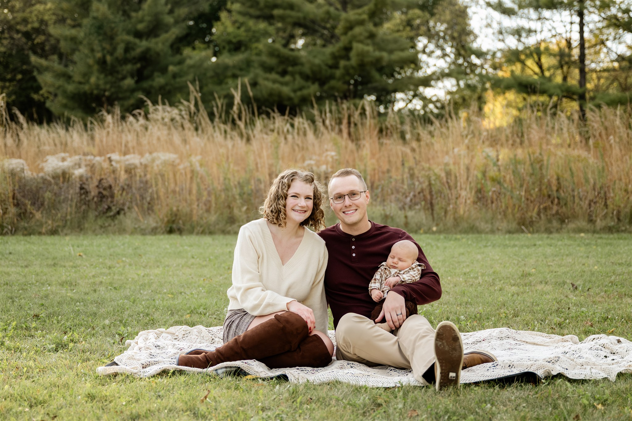 Happy mom and dad sit on a picnic blanket with their infant in dad's lap