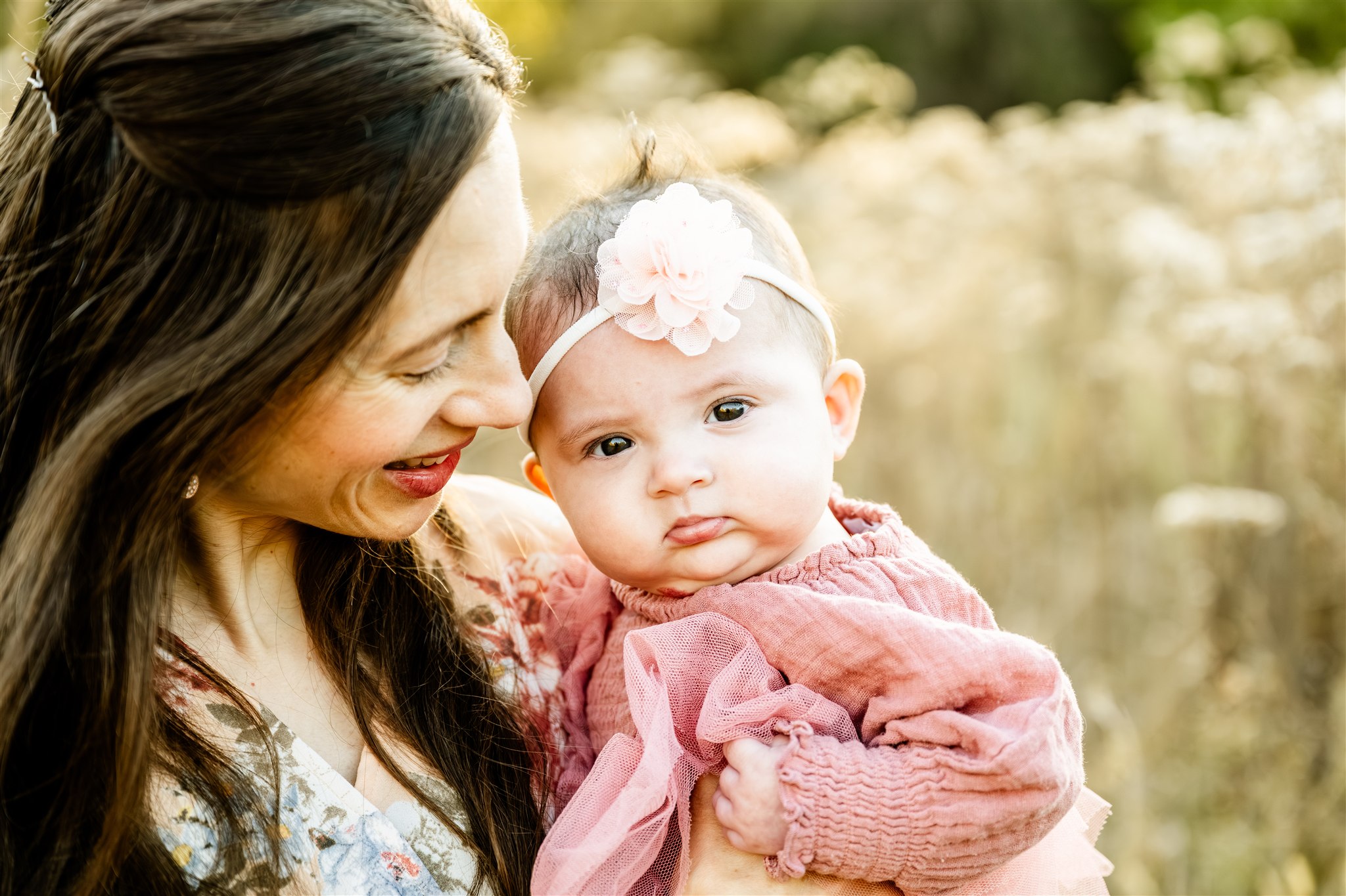 A happy mom snuggles her baby girl in a pink dress while standing in a park at sunset