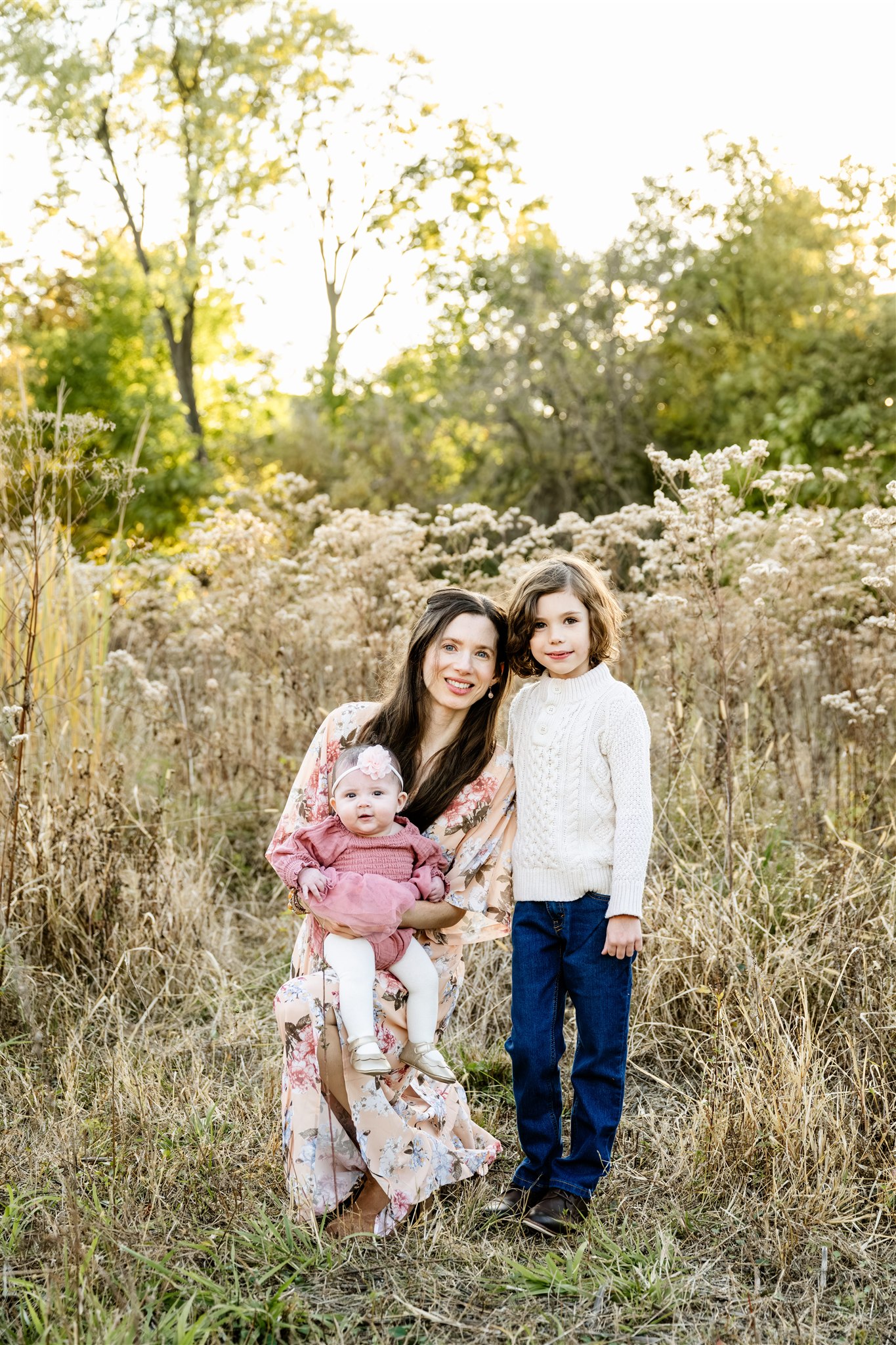 A happy mom in a floral print dress stands with her toddler son and baby girl in her lap in a park at sunset after visiting pediatricians in North Shore