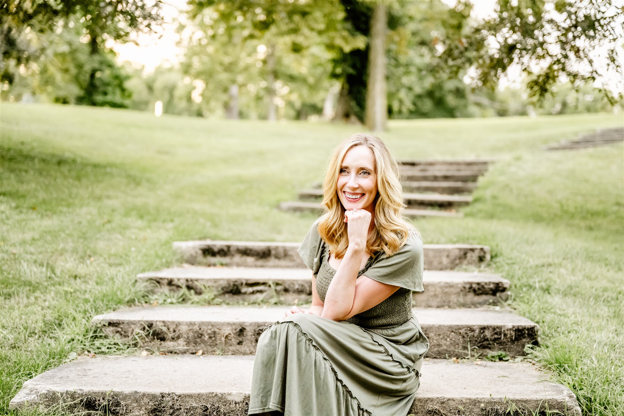 A smiling woman in a green dress sits on park steps leaning on her knee