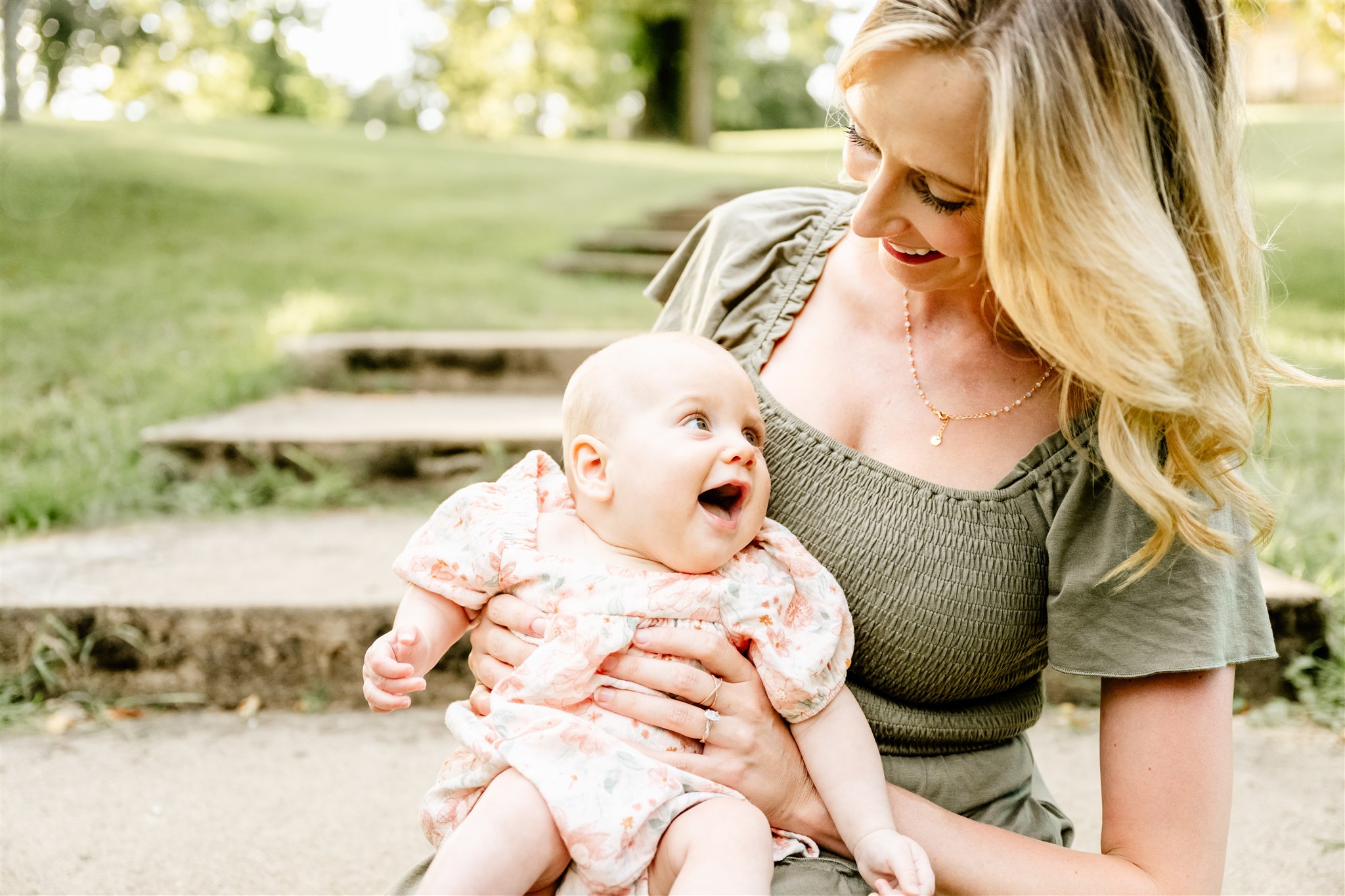 A happy mom sits on large park stairs playing with her infant daughter in her lap after some pilates classes in North Shore