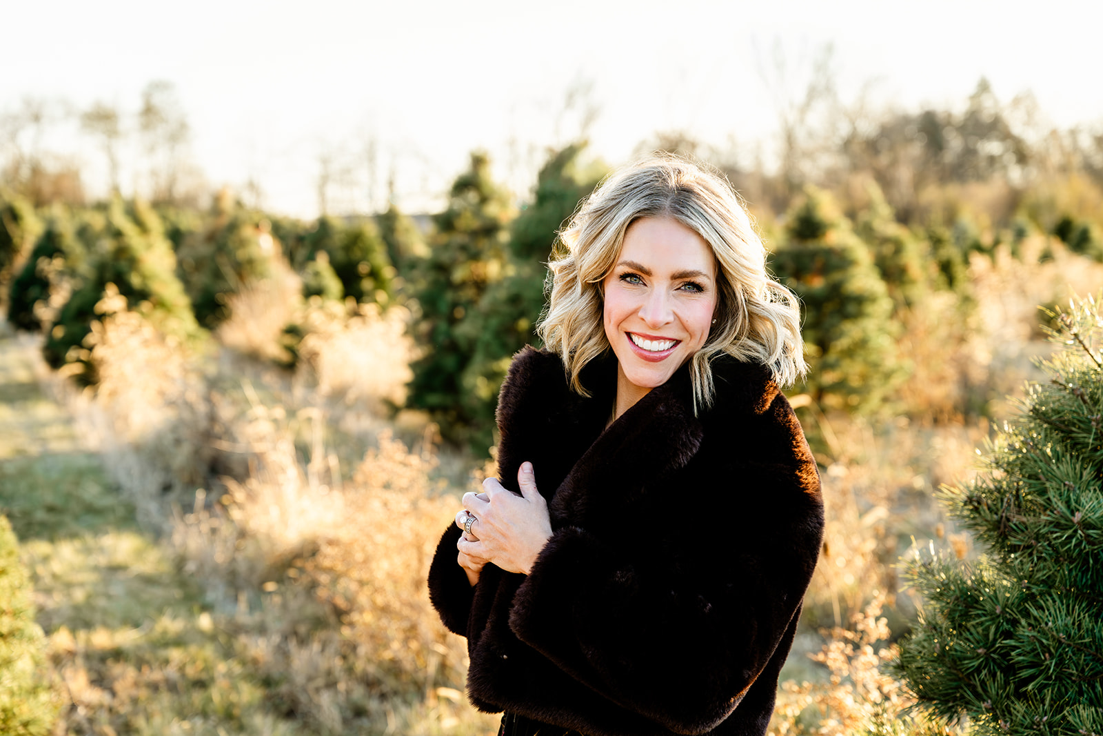 A woman smiles while standing in a christmas tree farm at sunset in a brown fur coat