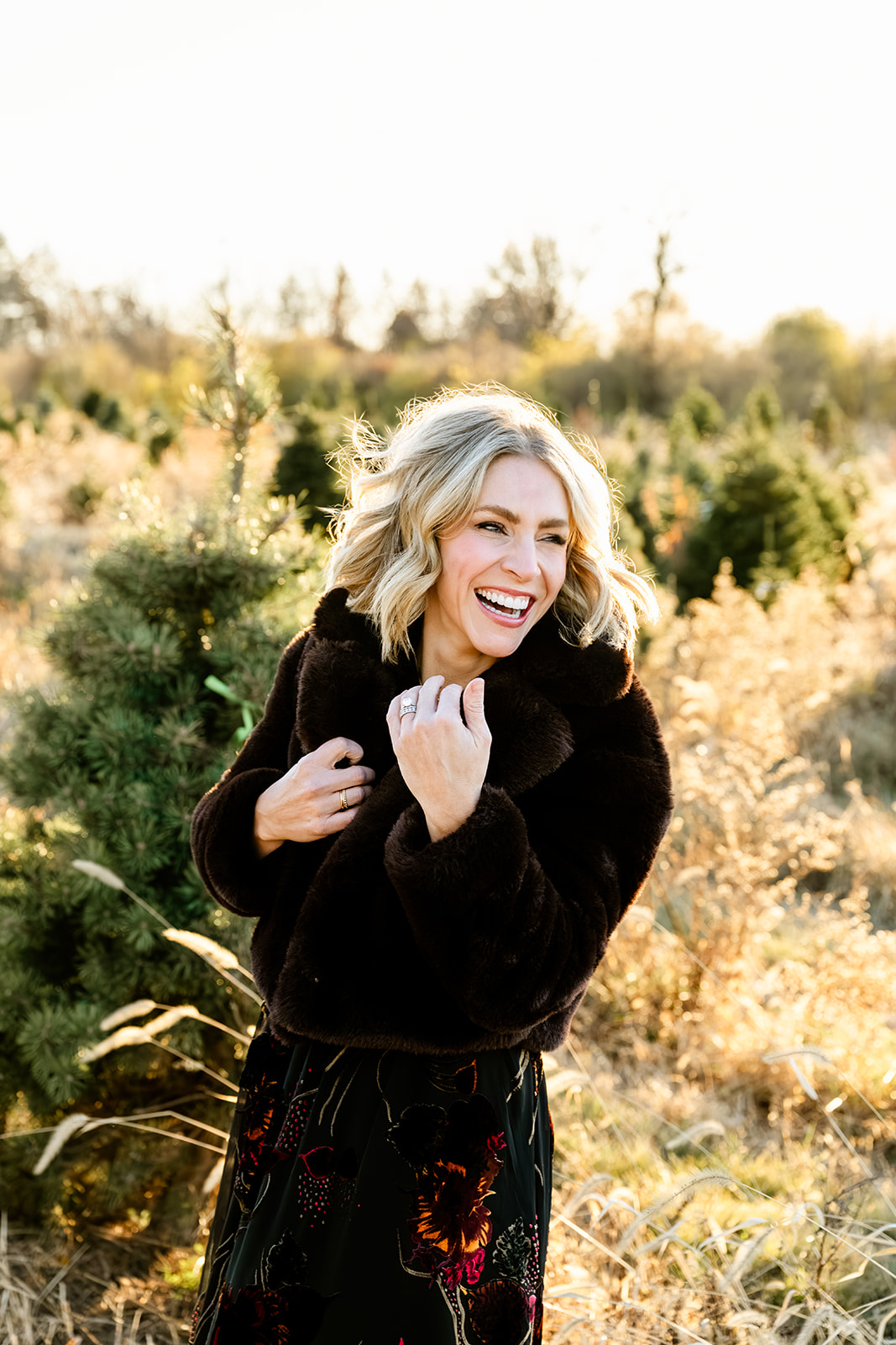 A woman laughs in a fur coat in a christmas tree farm at sunset after some pilates in Naperville