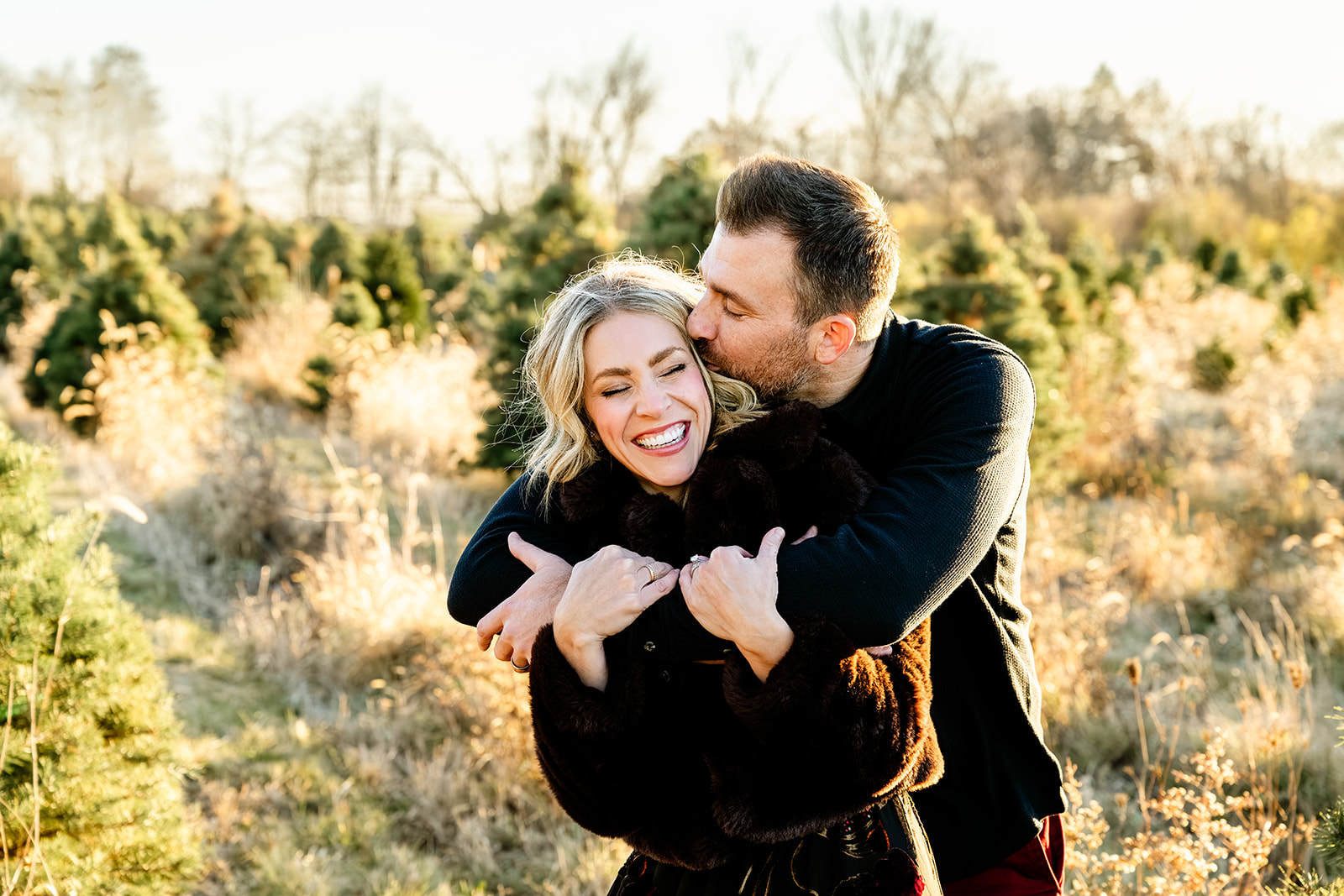 A man hugs and kisses the head of his smiling wife both in black sweaters in a park at sunset after some pilates in Naperville