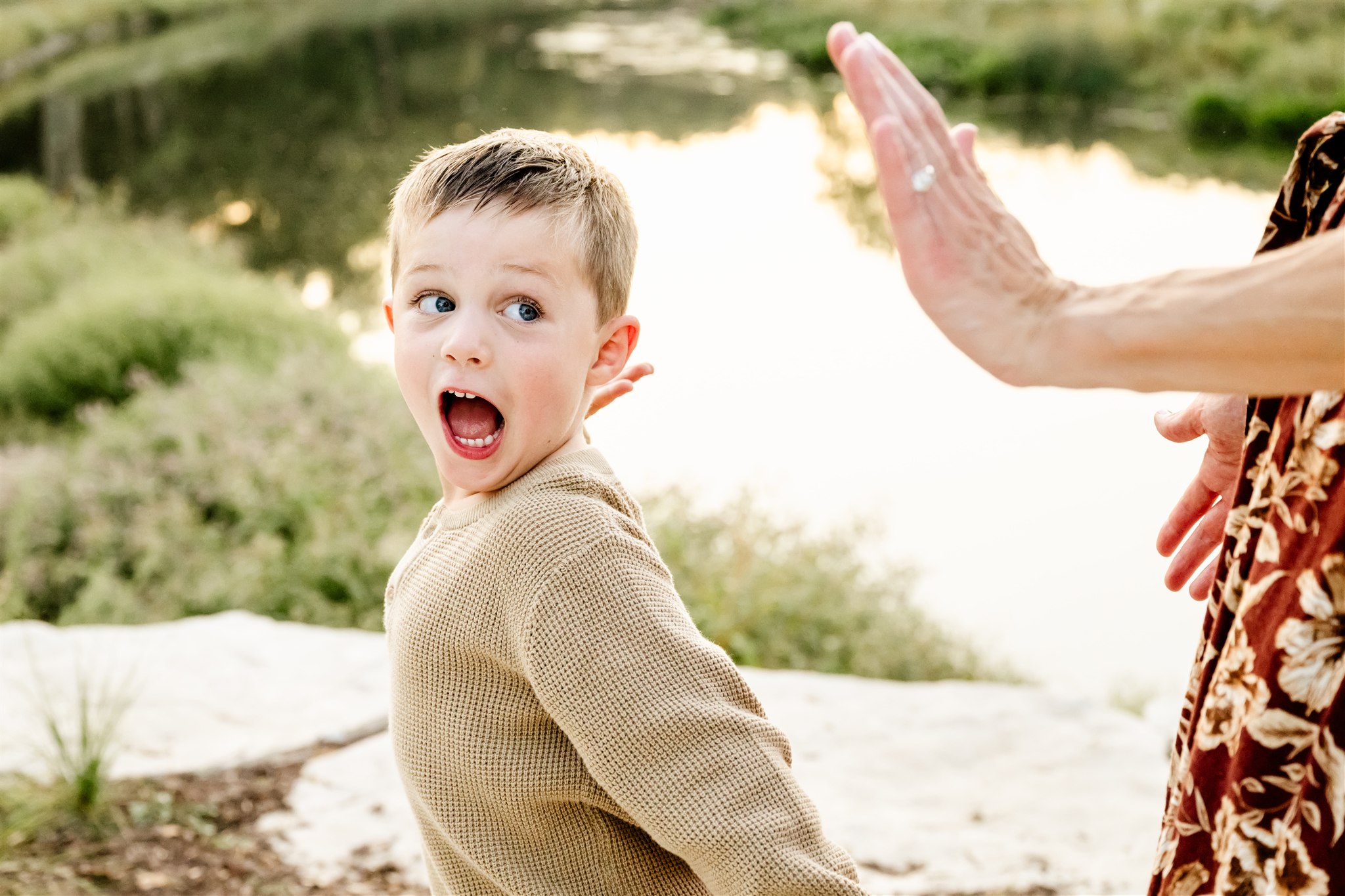 A toddler boy yells while rearing back for a high five to dad in a tan sweater by a pond at sunset
