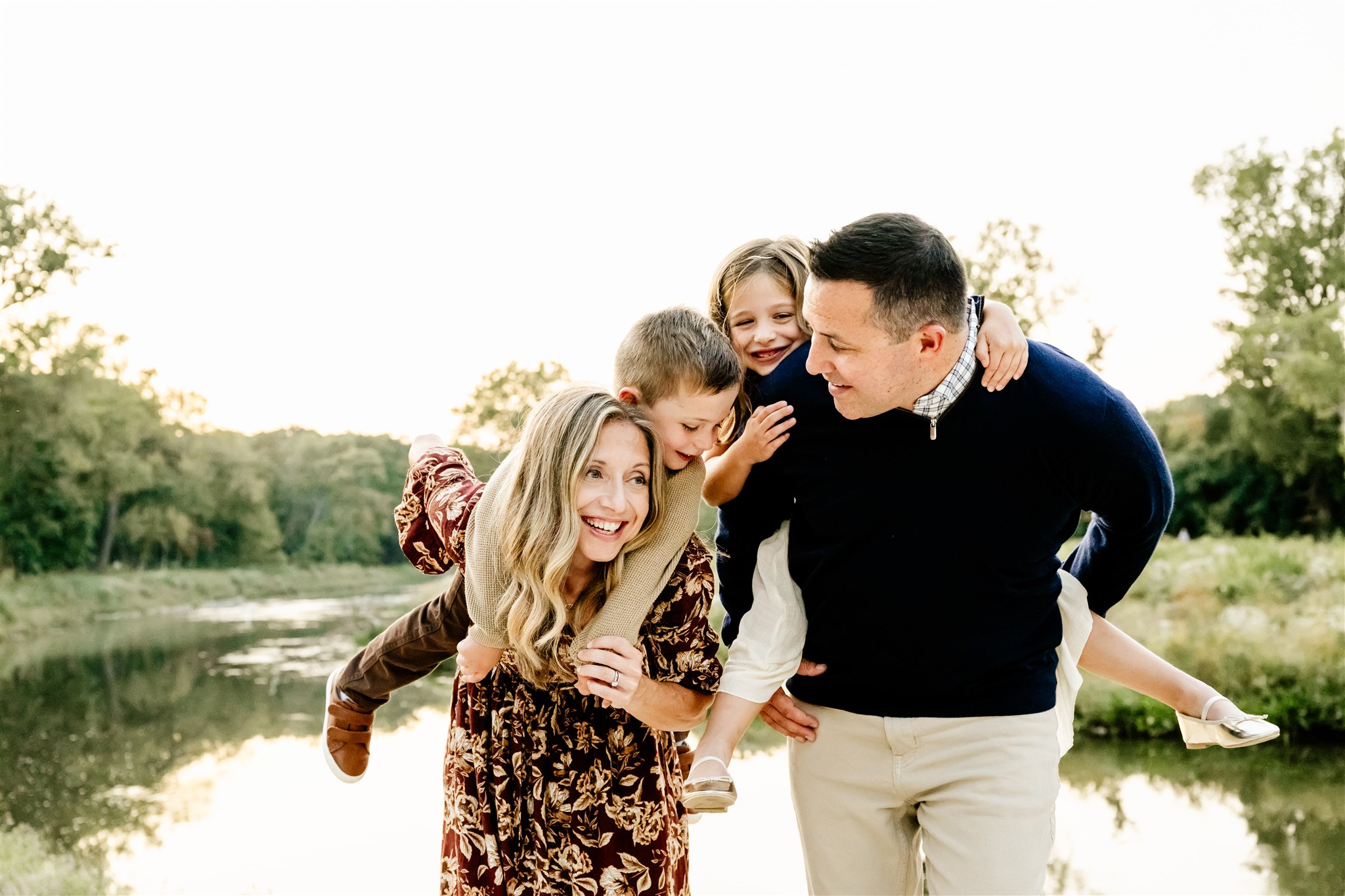 Mom and dad walk by a park pond with their toddler son and daughter on their backs at sunset after some play therapy in Naperville
