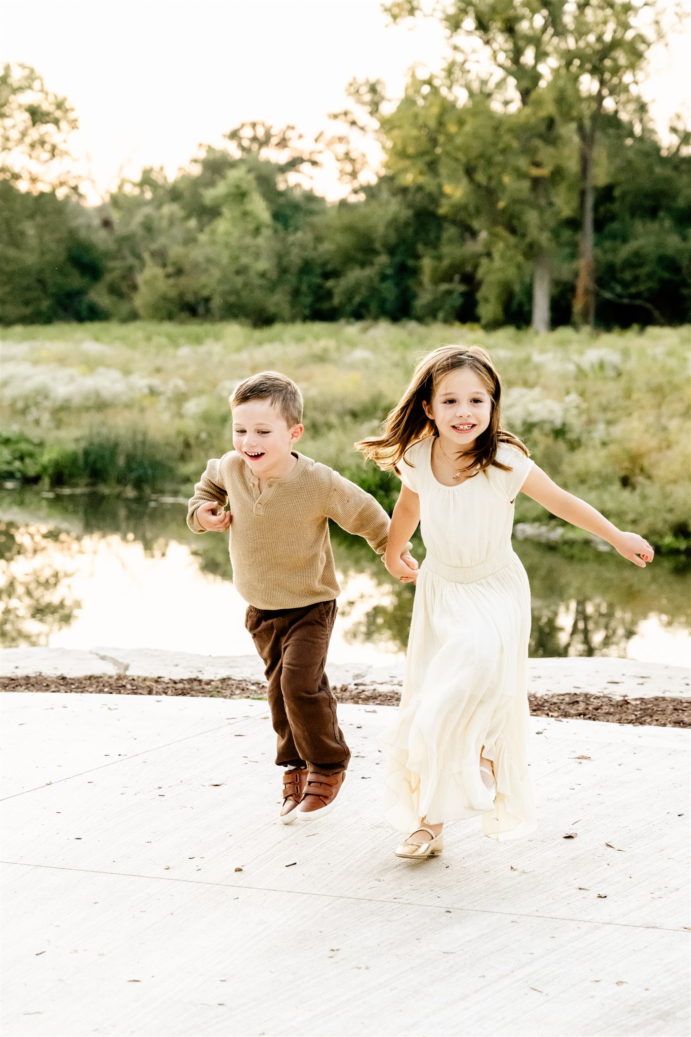 toddler brother and sister in tan and white hold hands while running by a pond at sunset thanks to play therapy in Naperville
