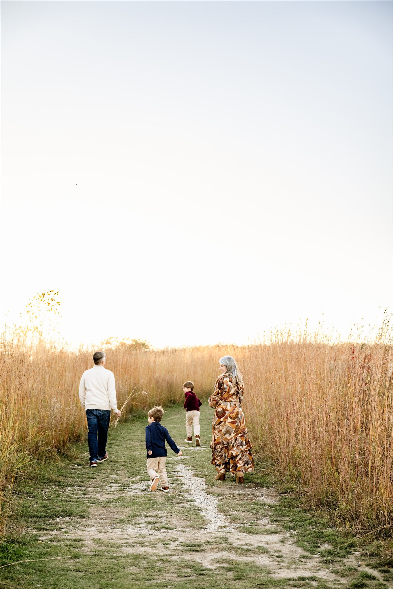A happy family with two toddler boys explores a path through tall golden grass at sunset