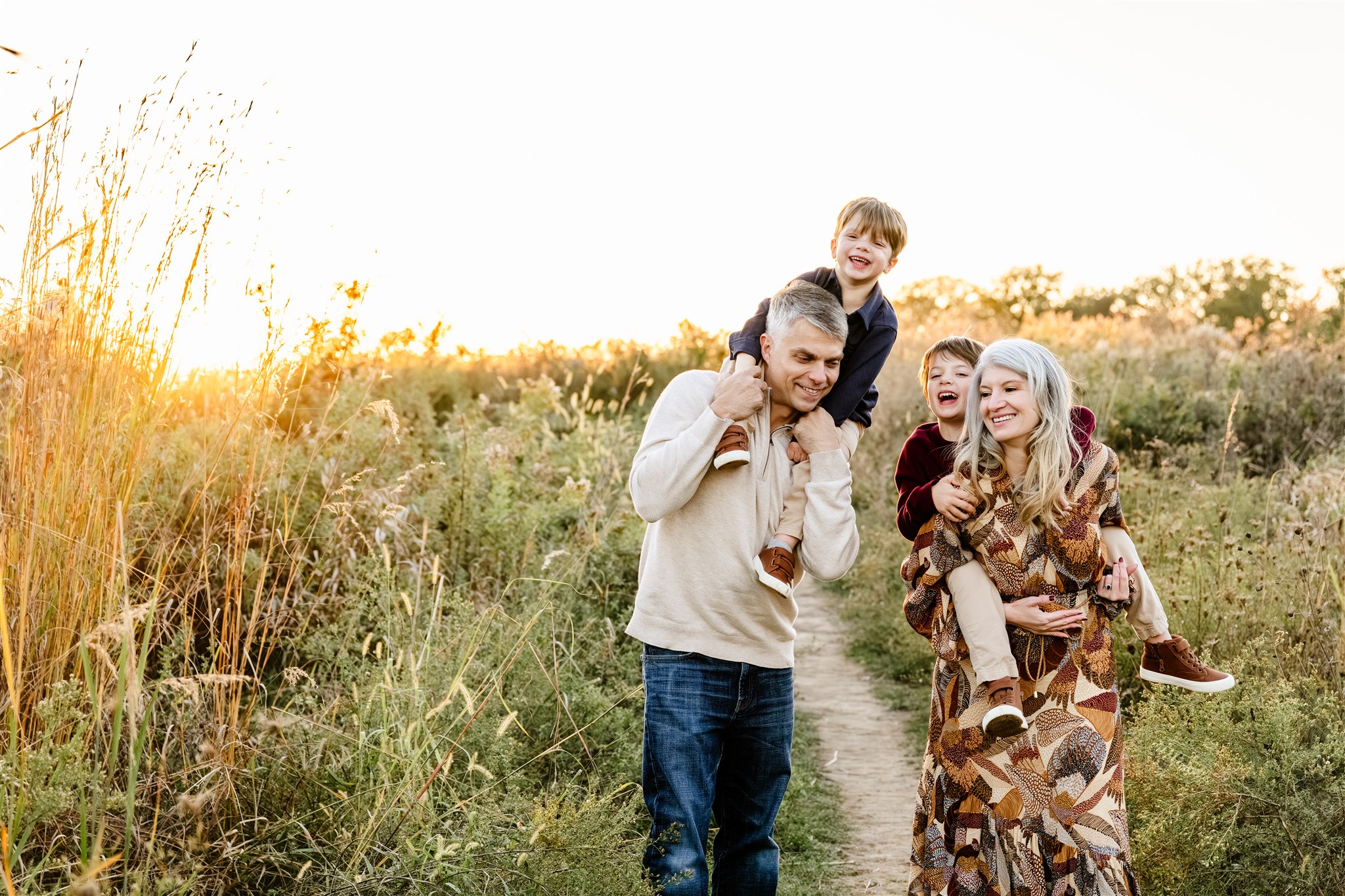 Happy toddler brothers ride on mom and dad's shoulders through a narrow trail at sunset in fall after visiting playgrounds in North Shore