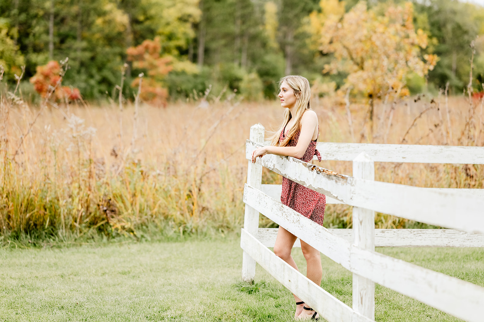 A high school senior leans on a pasture wooden fence at sunset in a red dress while smiling after enjoying private school in North Shore Chicago
