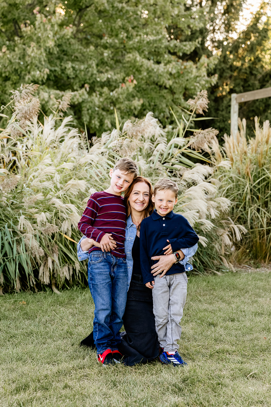 A happy mom in denim kneels to hug her smiling toddler sons in a garden at sunset after some speech therapy in Chicago