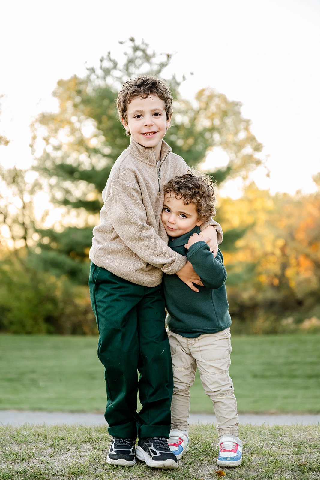 A young boy in tan hugs his little brother in a park in green at sunset after visiting toy stores in North Shore