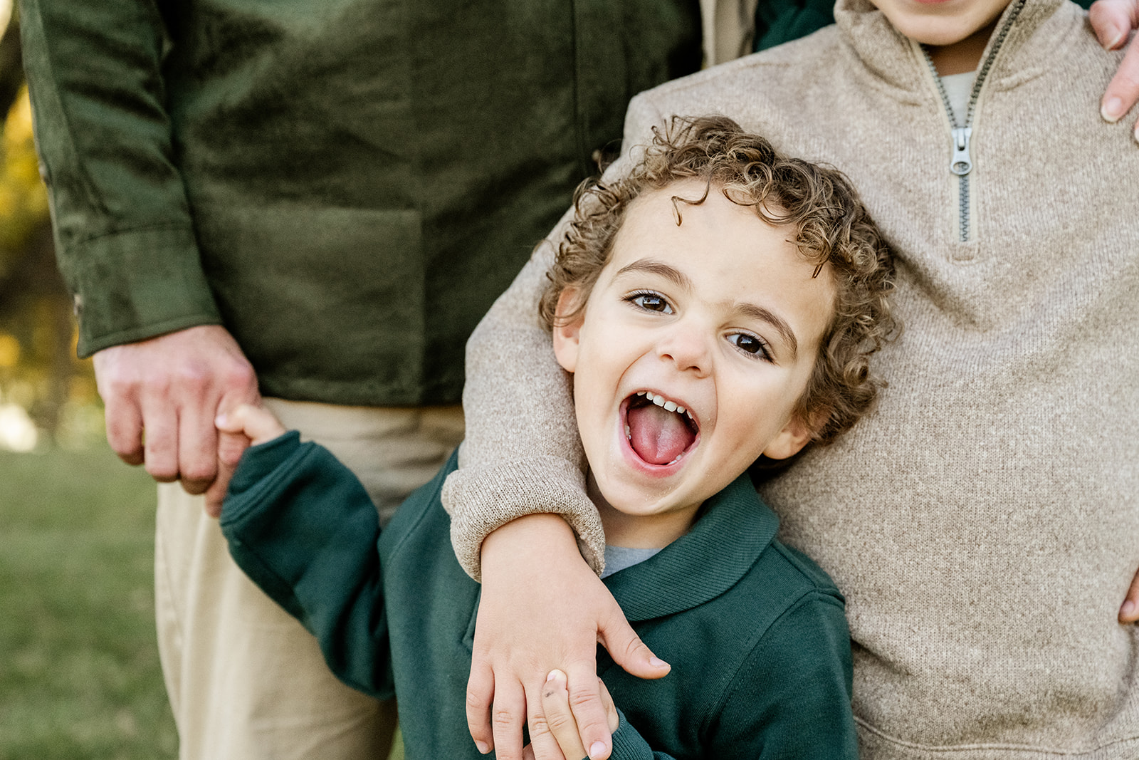 A happy toddler in a green sweater plays with big brother and dad in a park with big smiles after visiting toy stores in North Shore