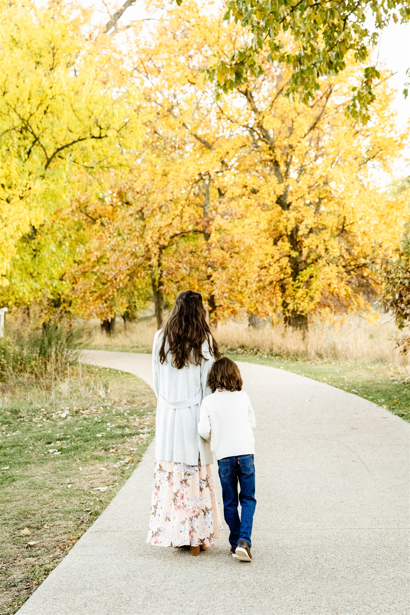 a mom walks in a park holding hands with her young son at sunset in fall