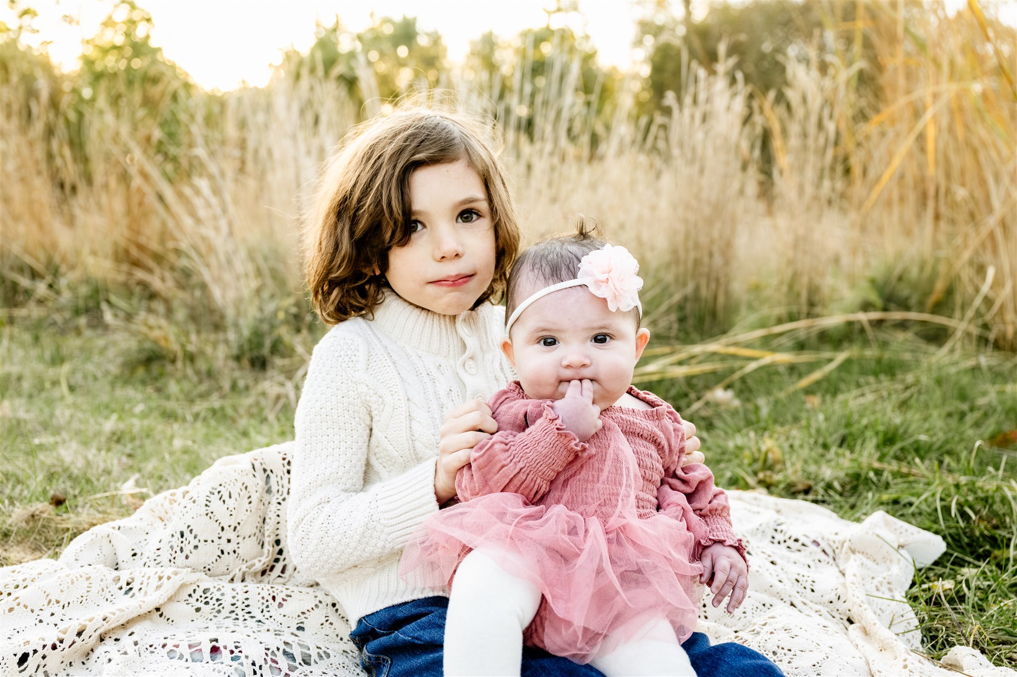 A young child in a white knit sweater sits with baby sister in pink in their lap on a lace picnic blanket in a park at sunset after exploring birthday party venues in North Shore Chicago