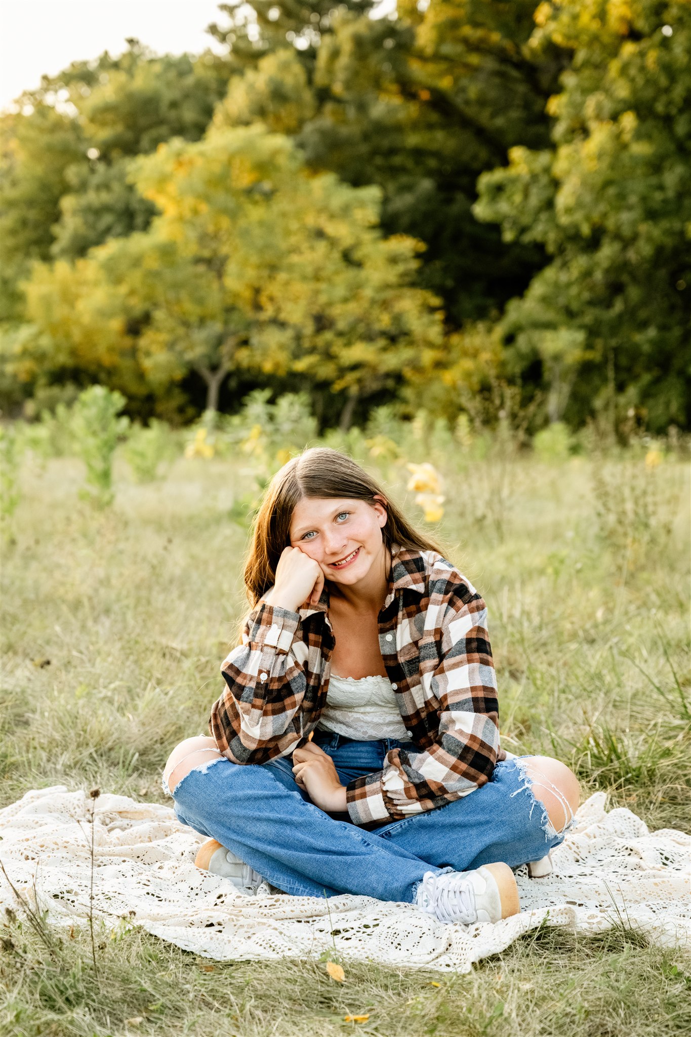 A smiling graduate sits on a lace picnic blanket in a park in plaid and jeans leaning on her knee