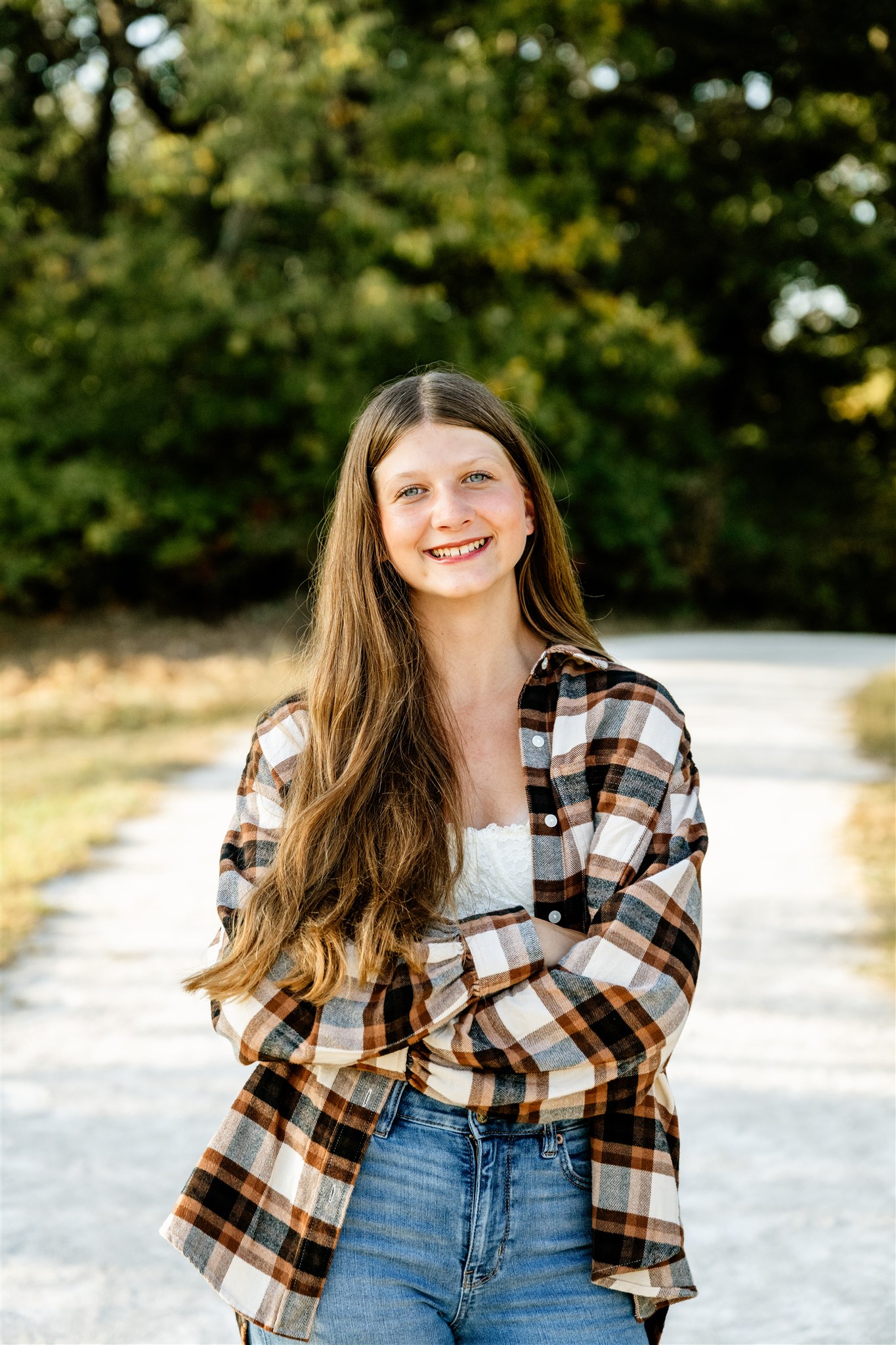 A happy high school senior in a plaid shirt and jeans stands in a park sidewalk with arms crossed after visiting boutiques in North Shore