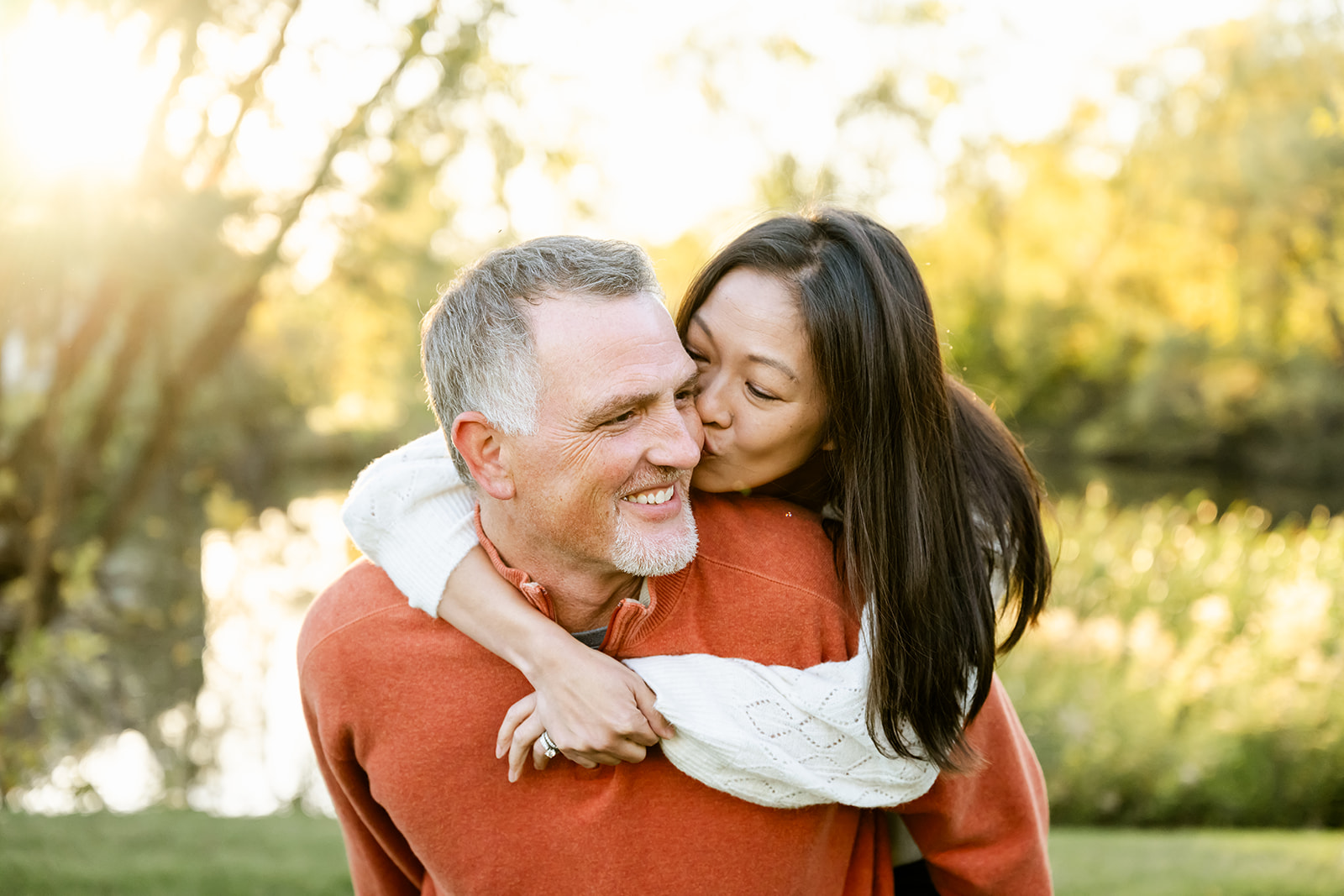 A man in an orange sweater is kissed on the cheek by his wife on his back in white by a pond at sunset