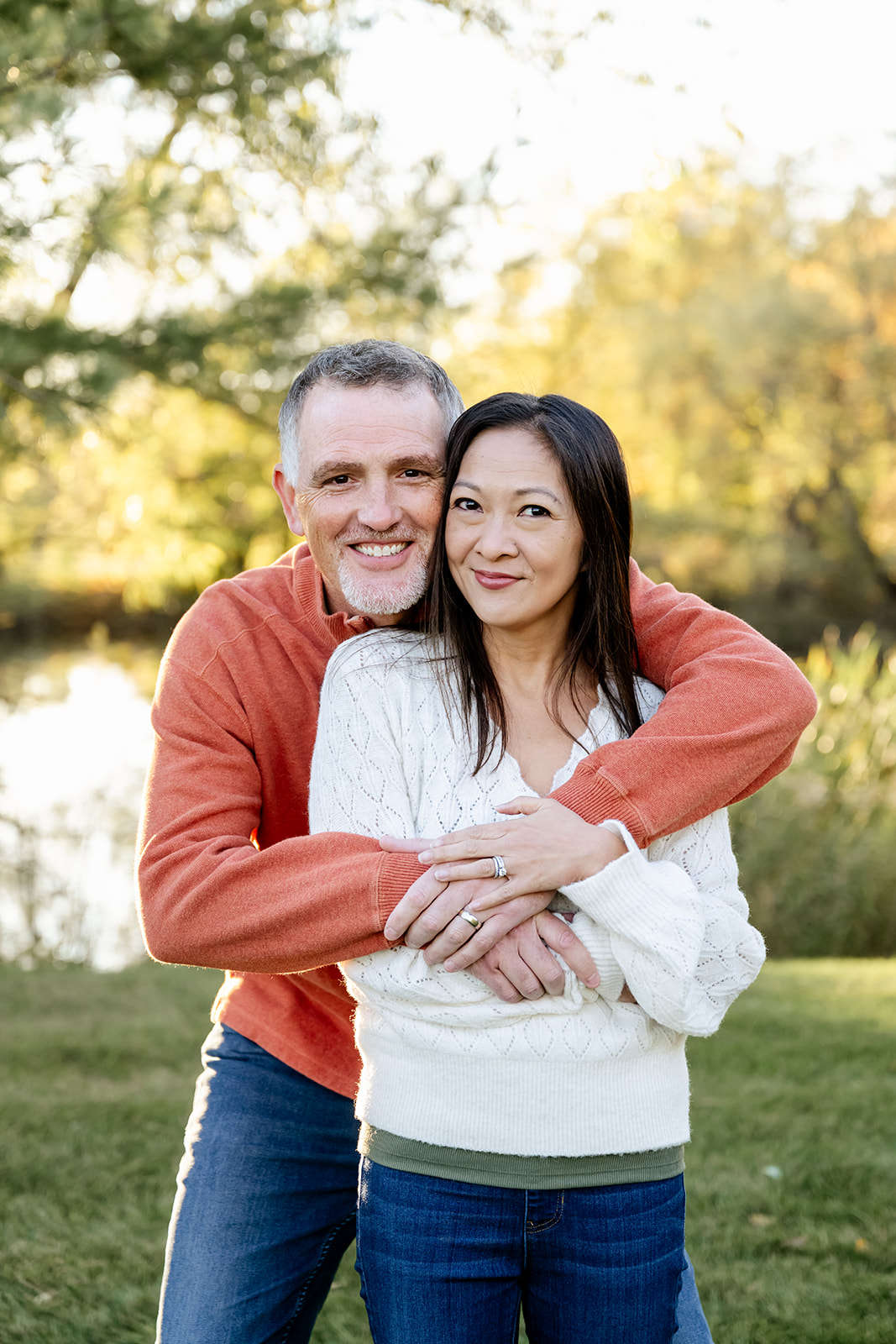 A happy man in an orange sweater hugs his wife from behind in a park while enjoying date night ideas in Naperville
