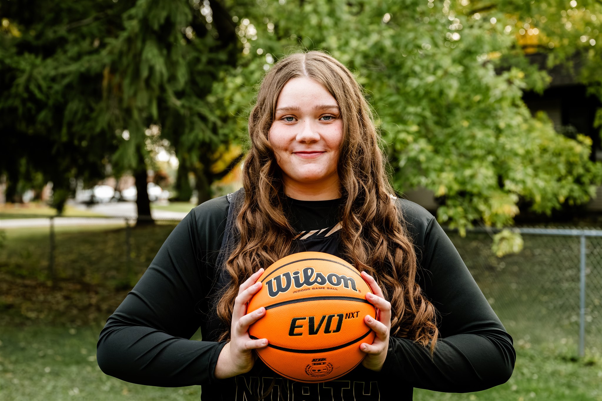 A happy high school basketball player holds her ball while standing in a lawn smiling in her black uniform after enjoying hair salons in North Shore Chicago