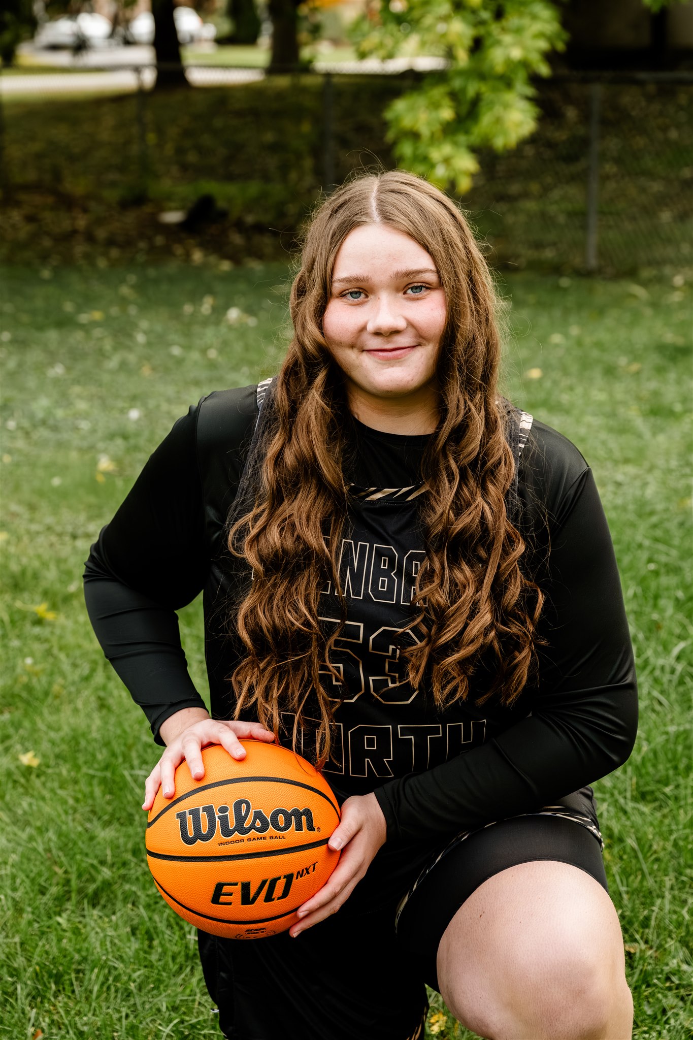 A high school basketball player kneels in a lawn in her balck uniform with a ball after finding great hair salons in North Shore Chicago
