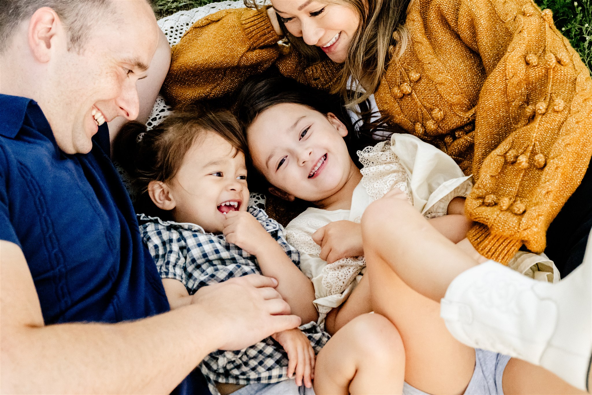Toddler sisters are tickled by mom and dad while laying on a picnic blanket