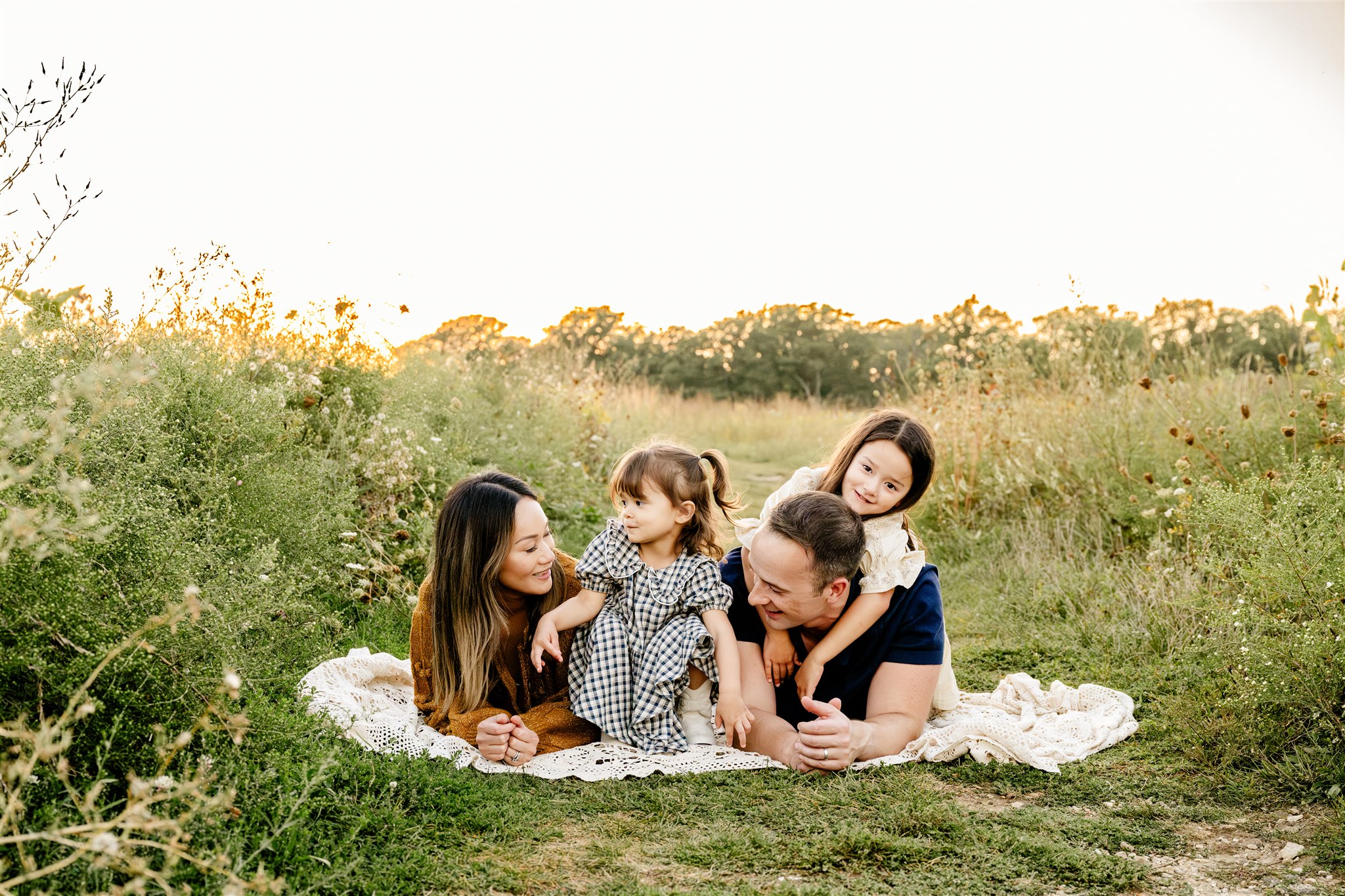 A mom and dad lay on a picnic blanket with their toddler daughters crawling on them in dresses at sunset in a park trail before visiting ice cream shops in Chicago