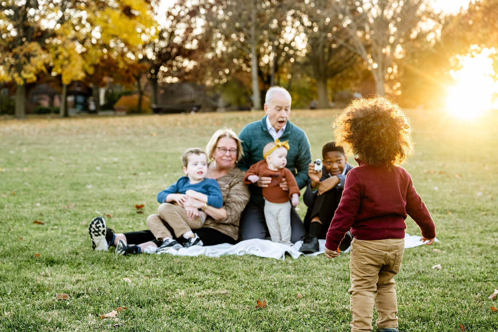Grandma and grandpa on a picnic blanket with three grandkids in their laps as another walks towards them at sunset