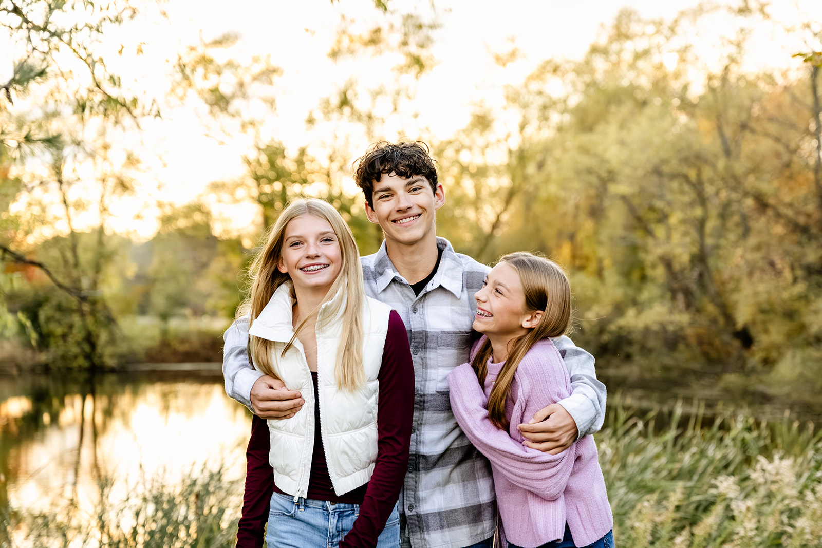 A brother hugs his two sisters on each side of him as they smile and stand in front of a pond with surrounding trees at sunset.