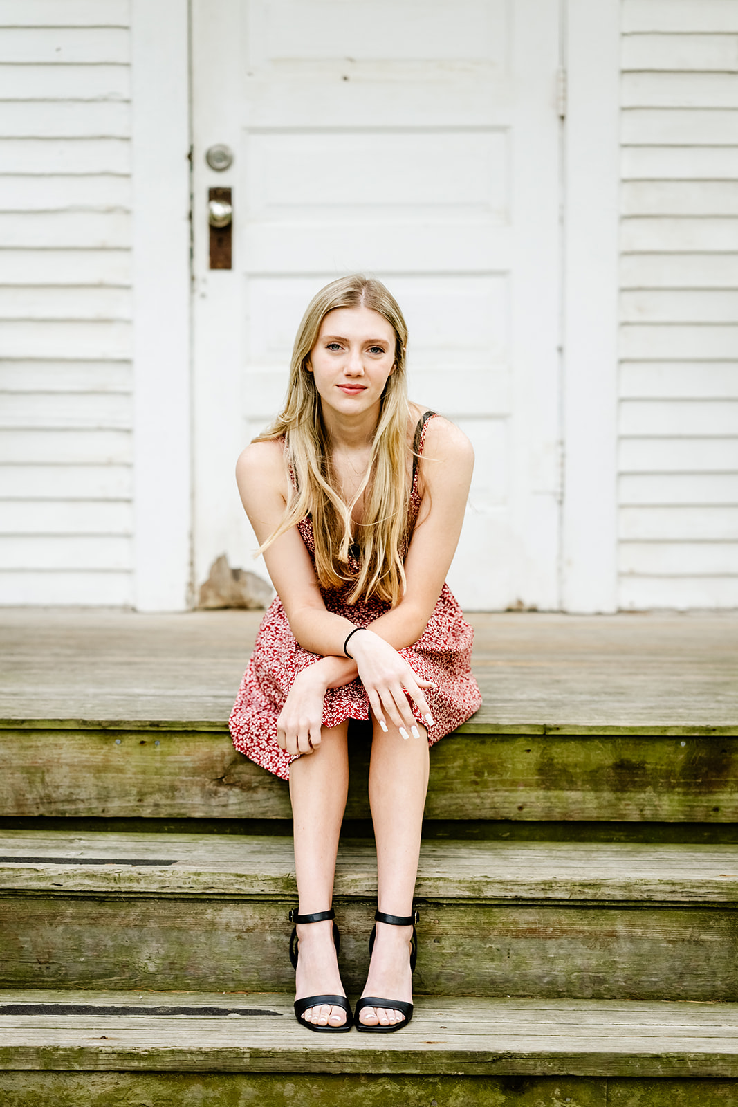 A high school senior sits on an old wooden stoop leaning on her knees in a red dress
