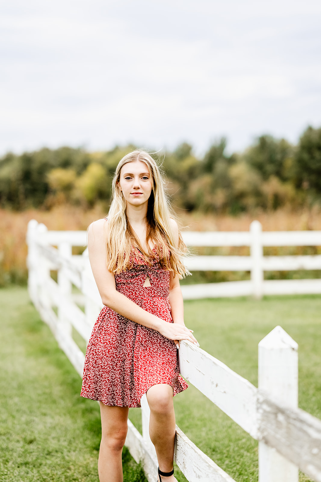 A high school senior leans on a wooden fence in a park lawn in a red dress after visiting nail salons in North Shore Chicago