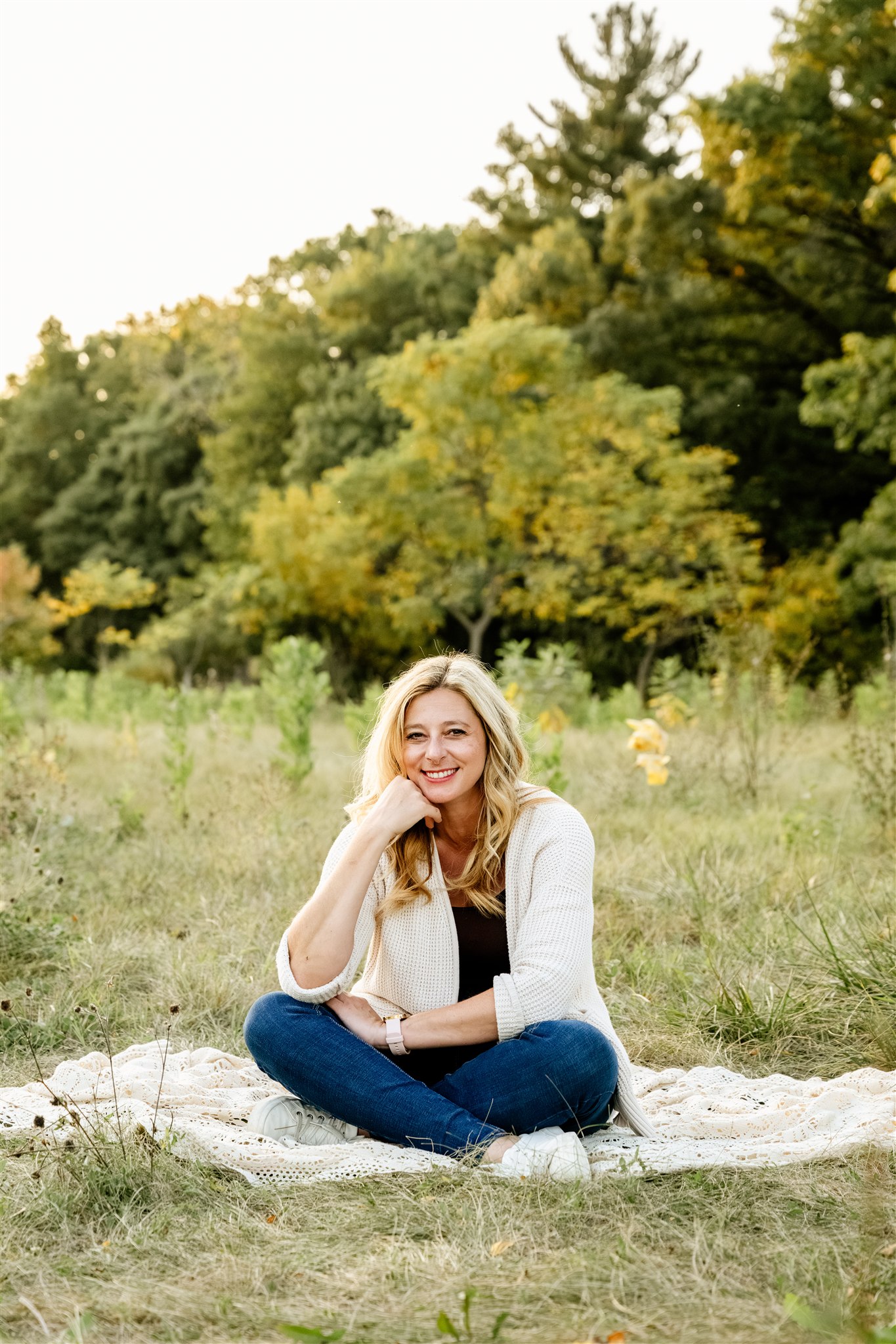 A lady smiles as she is sitting on a blanket in a field of grass wearing jeans and a sweater,