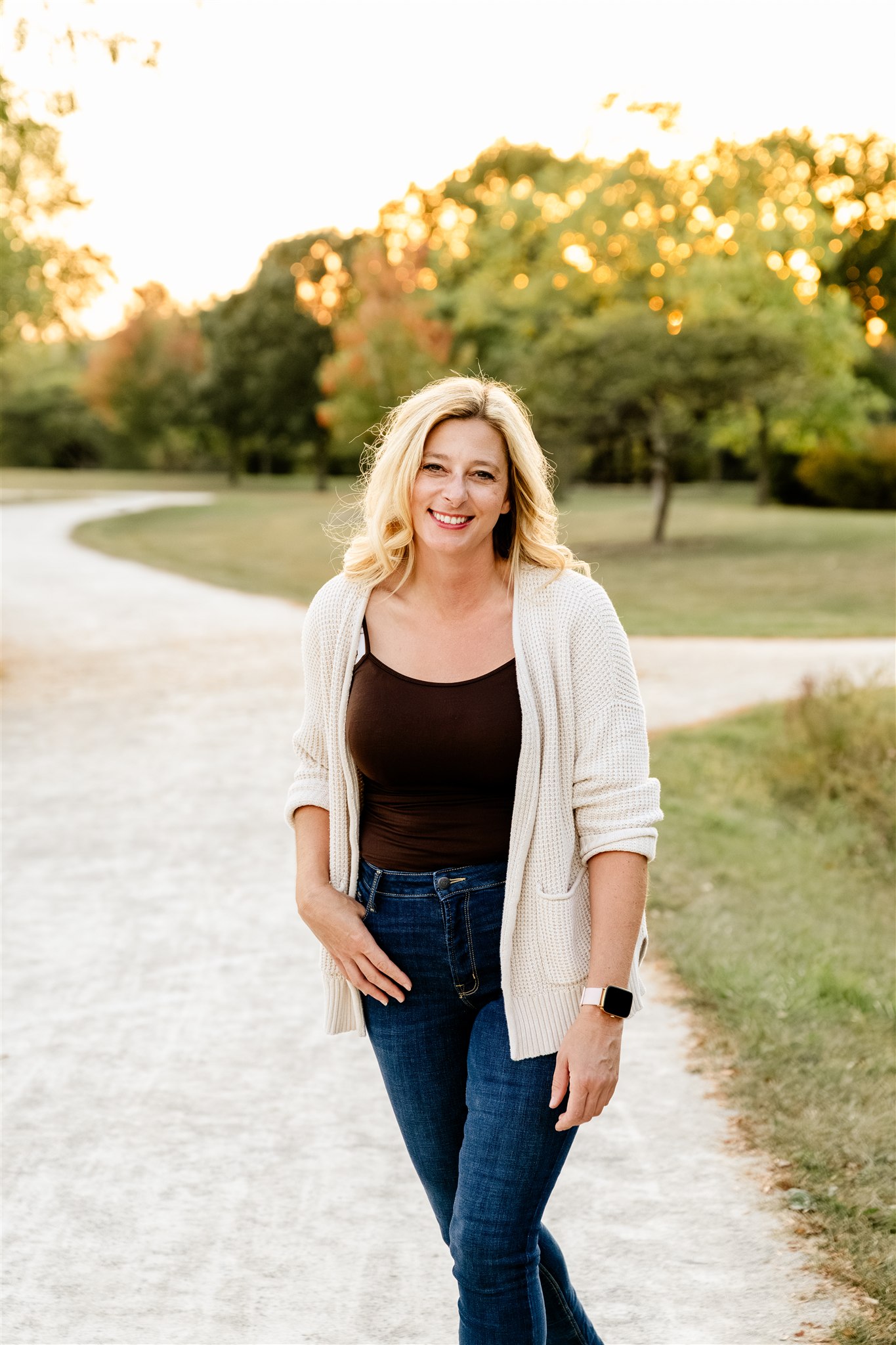 A lady in a brown top and jeans with a white sweater smiles and stands in a park after a pilates class in chicago.