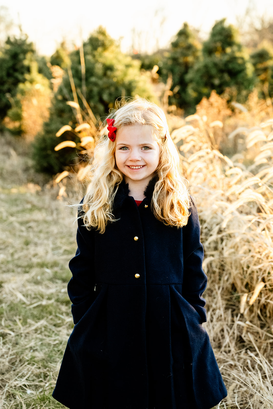 A young girl in a black coat stands smiling with hands in her pockets in a christmas tree farm after visiting preschools in North Shore
